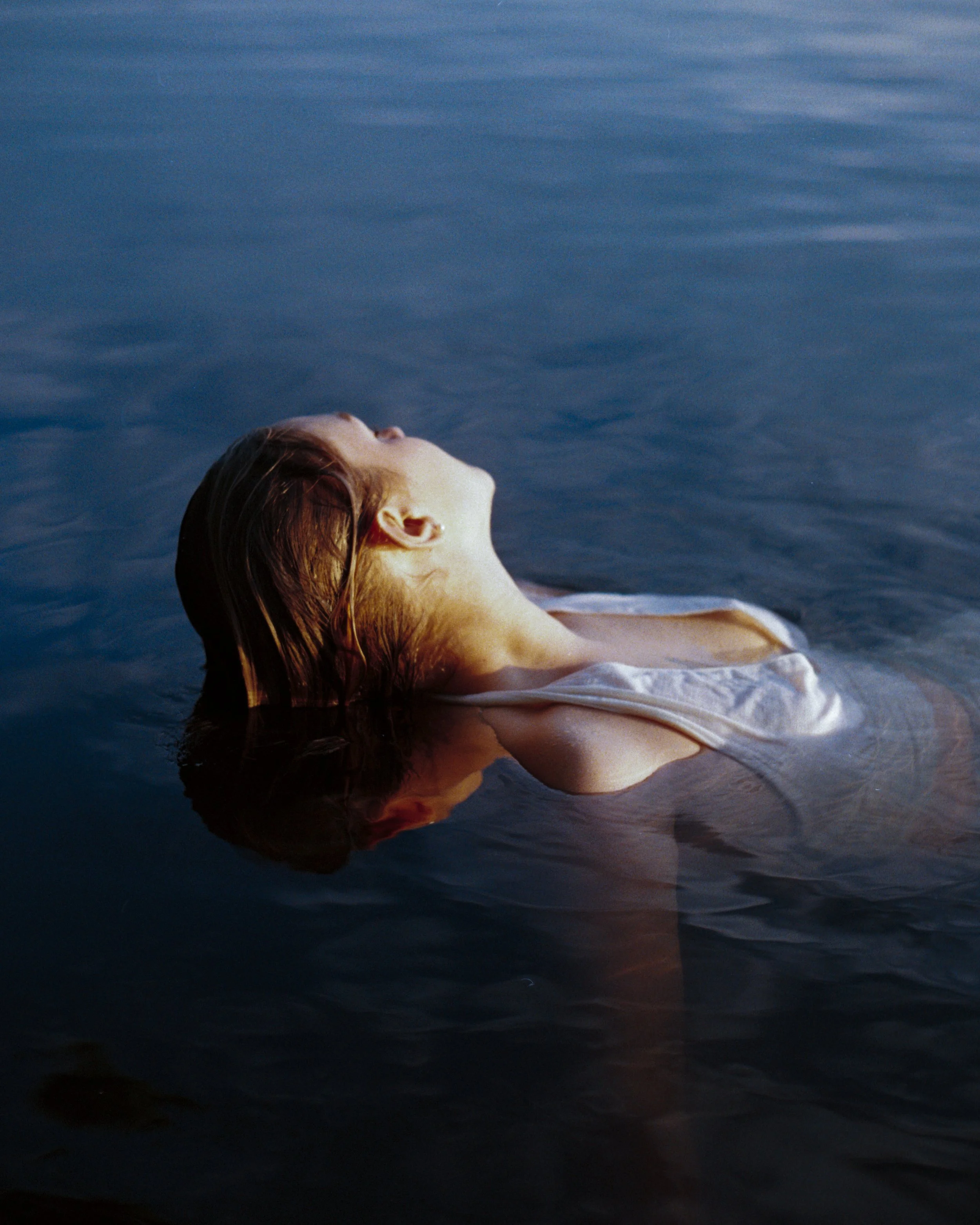 A woman with wet hair lying in shallow water with her head tilted back and eyes closed, at sunset or sunrise.