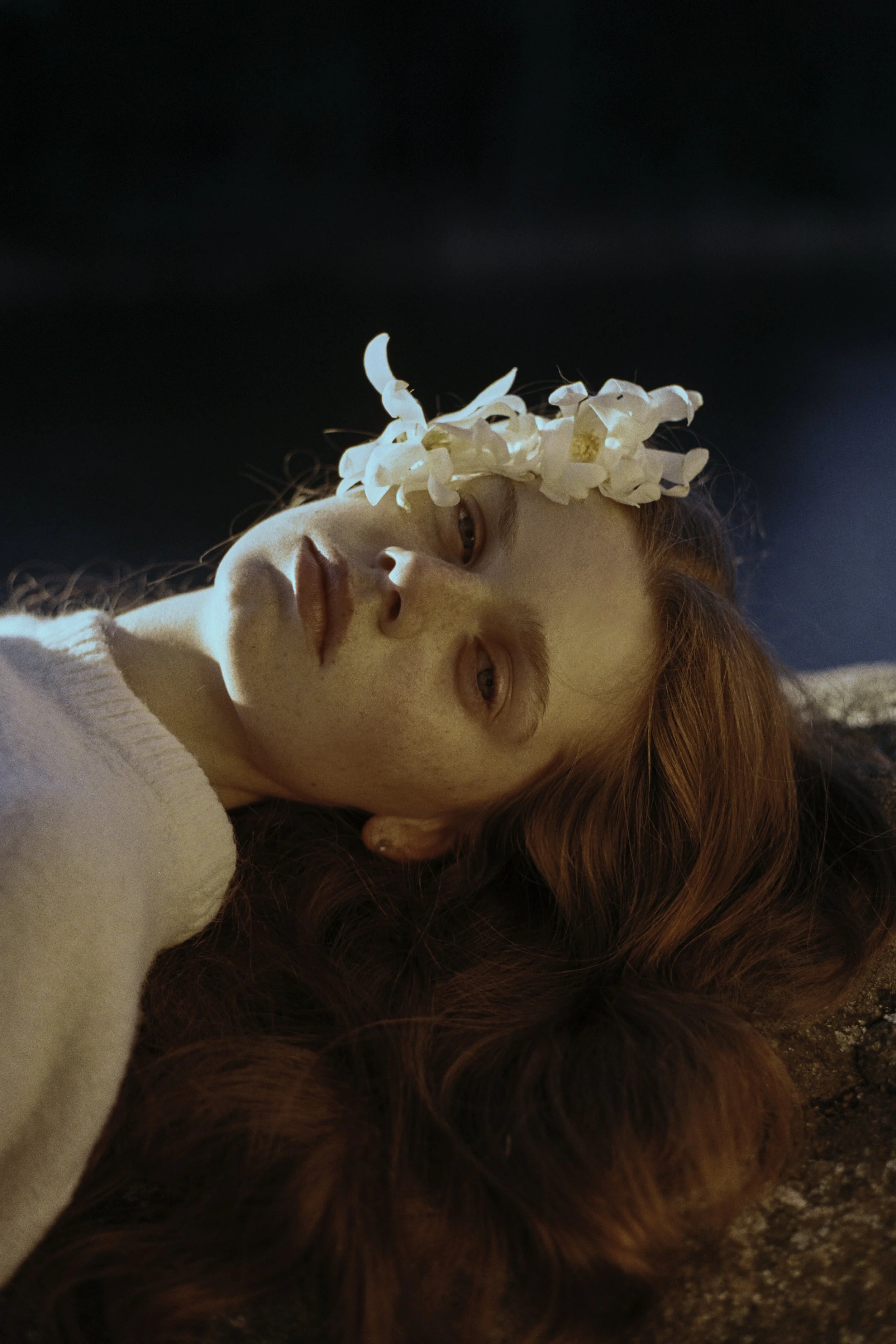 A woman with red hair lying down on a textured surface, wearing a floral crown.
