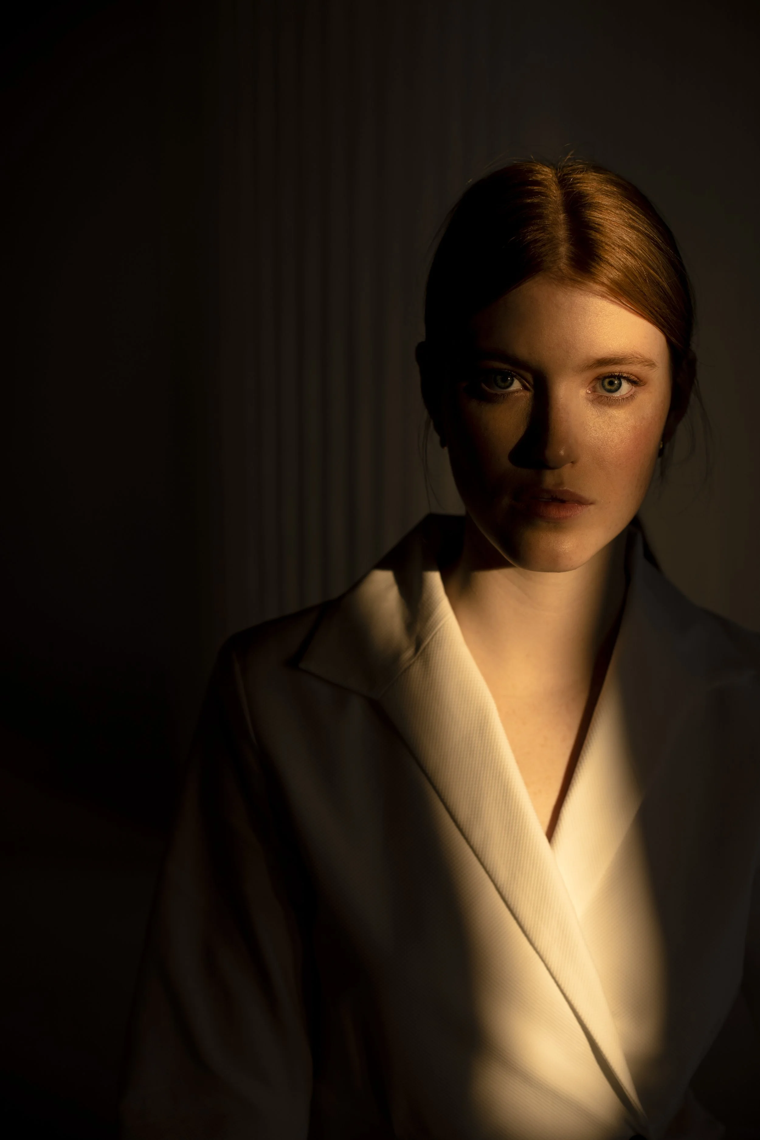 Portrait of a young woman with red hair and piercing blue eyes, wearing a beige blazer, illuminated by soft lighting against a dark background.
