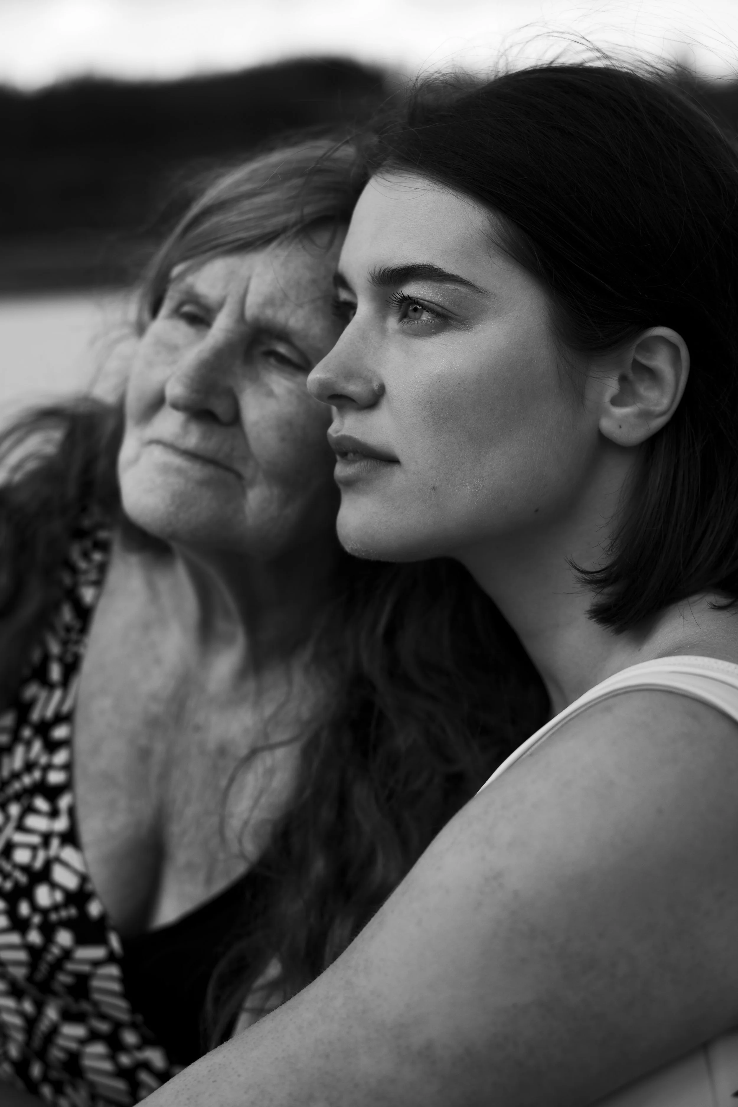 Black and white photograph of an elderly woman with closed eyes and a young woman with open eyes and a slight smile resting their heads together outdoors.