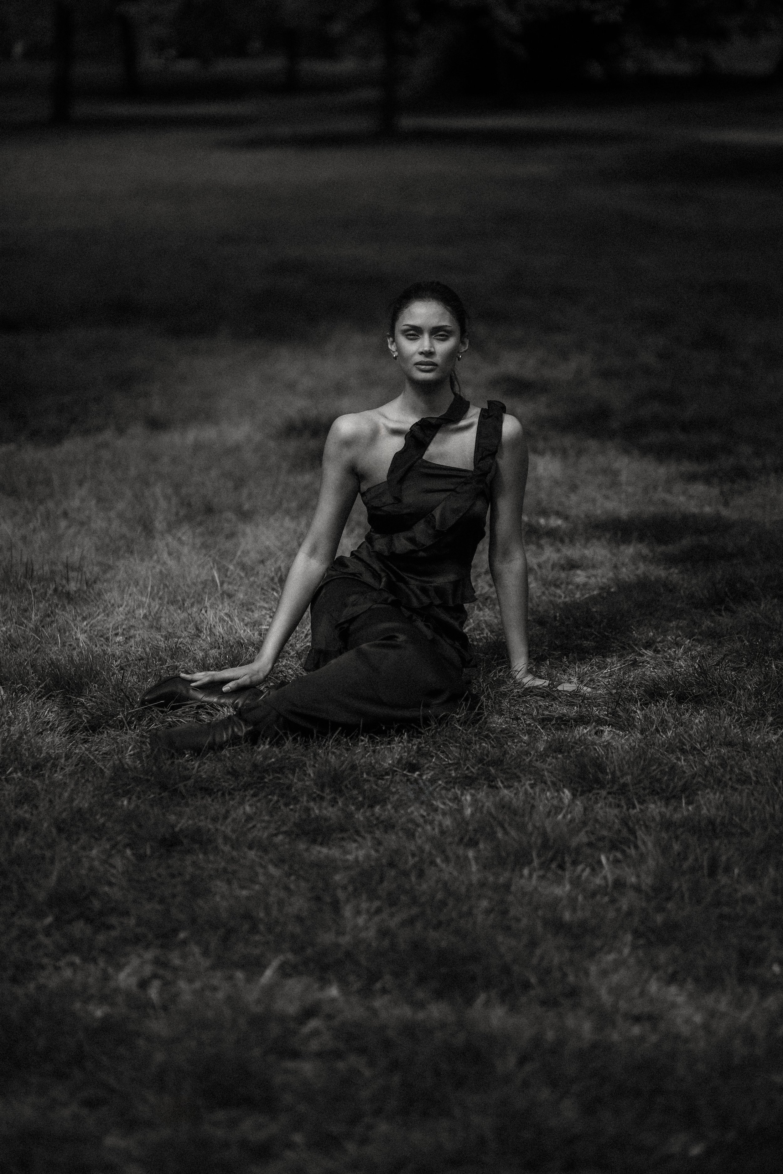 Black and white photo of a woman sitting on the grass outdoors at night, wearing a dress with ruffles.