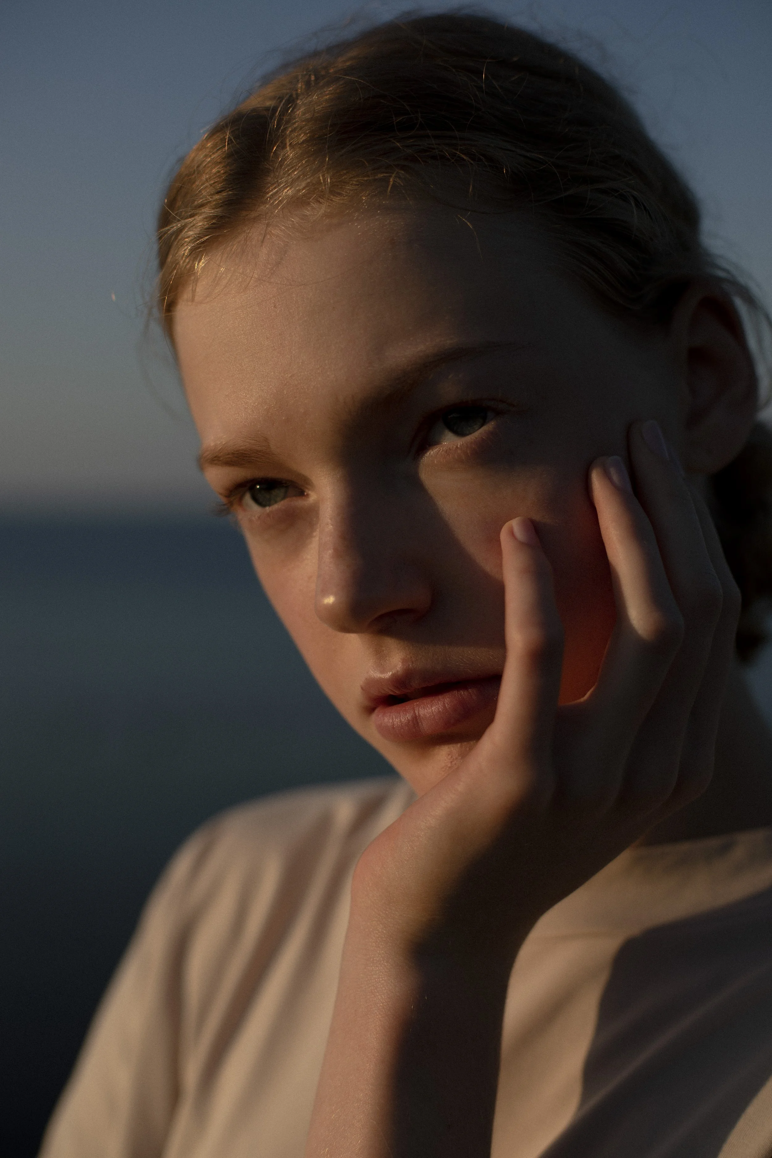 A young woman with blonde hair and blue eyes touching her face with her fingers, looking into the distance in soft lighting.