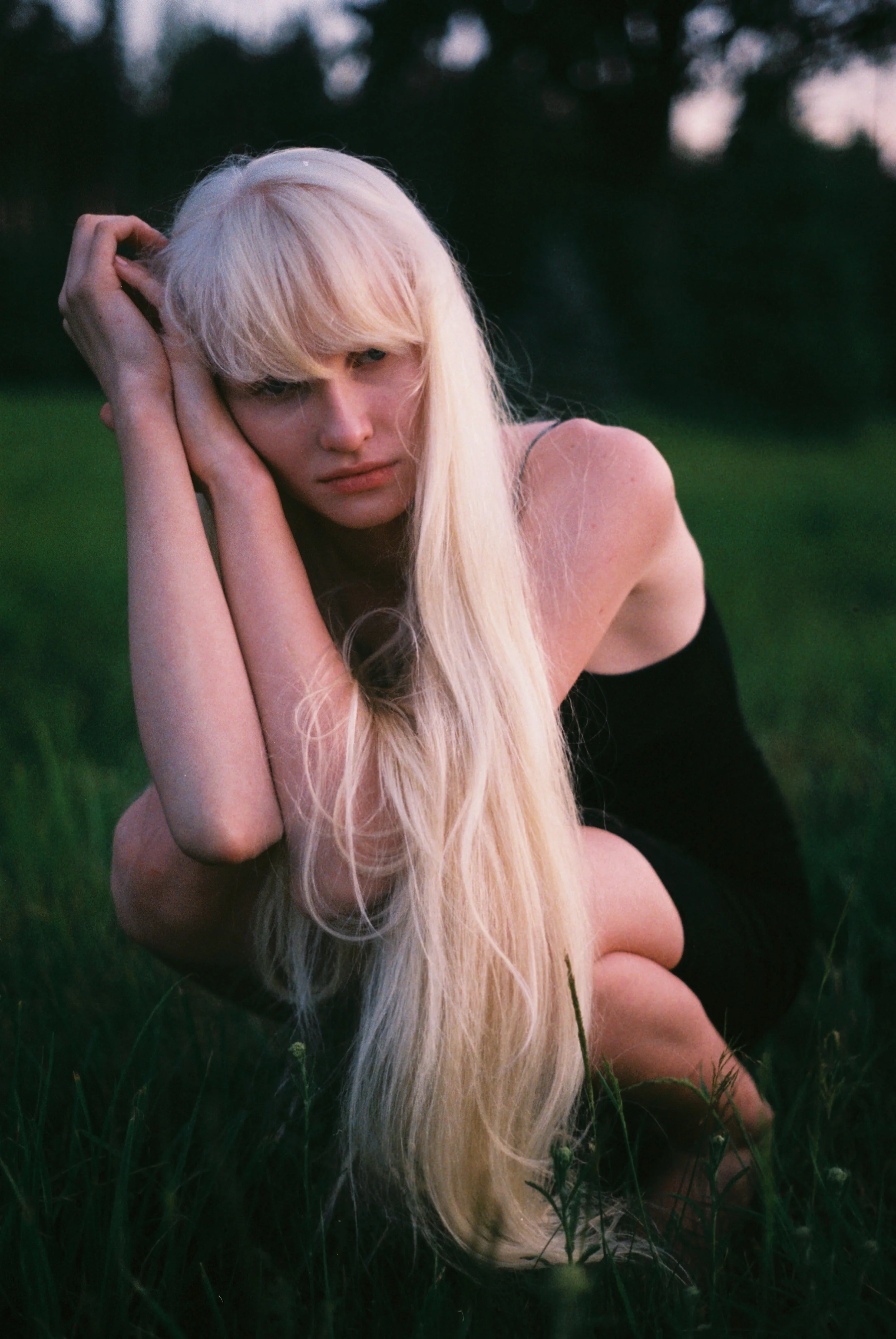 A woman with long blonde hair poses outdoors on grass at dusk, resting her head on her hands with a somber expression.