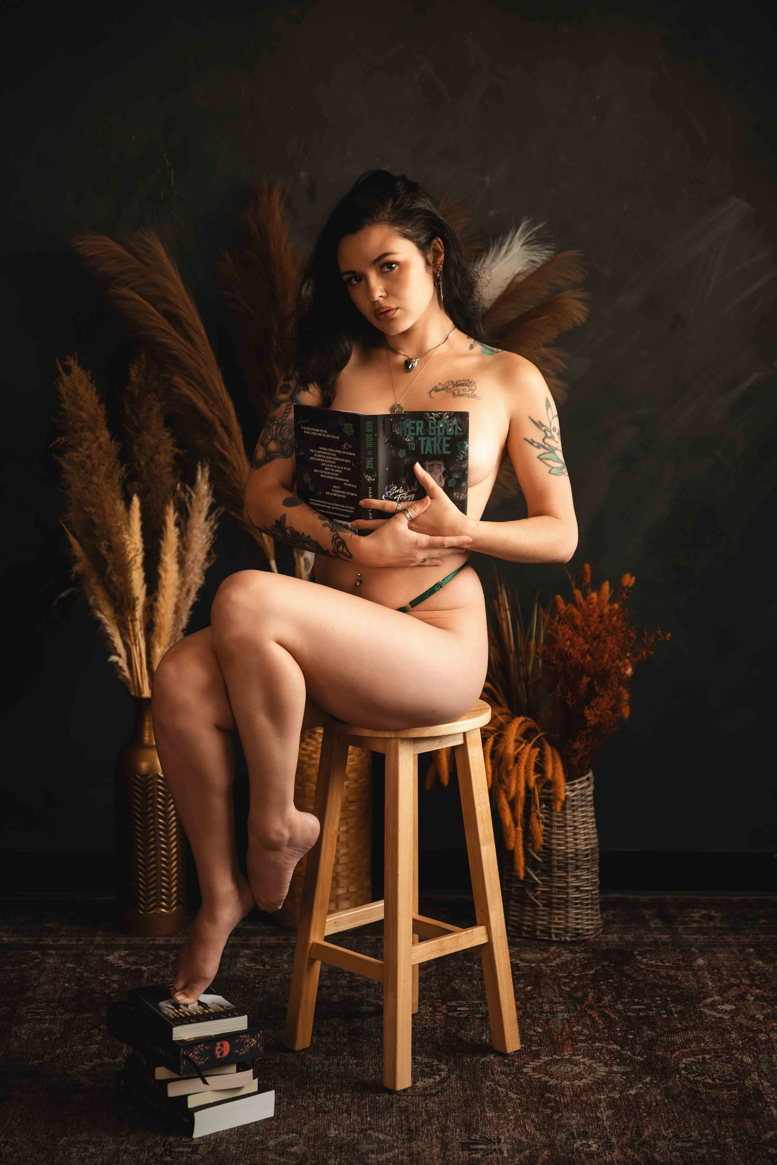 Tattooed woman seated on a stool holding a book during a dark botanical book-themed Houston boudoir session