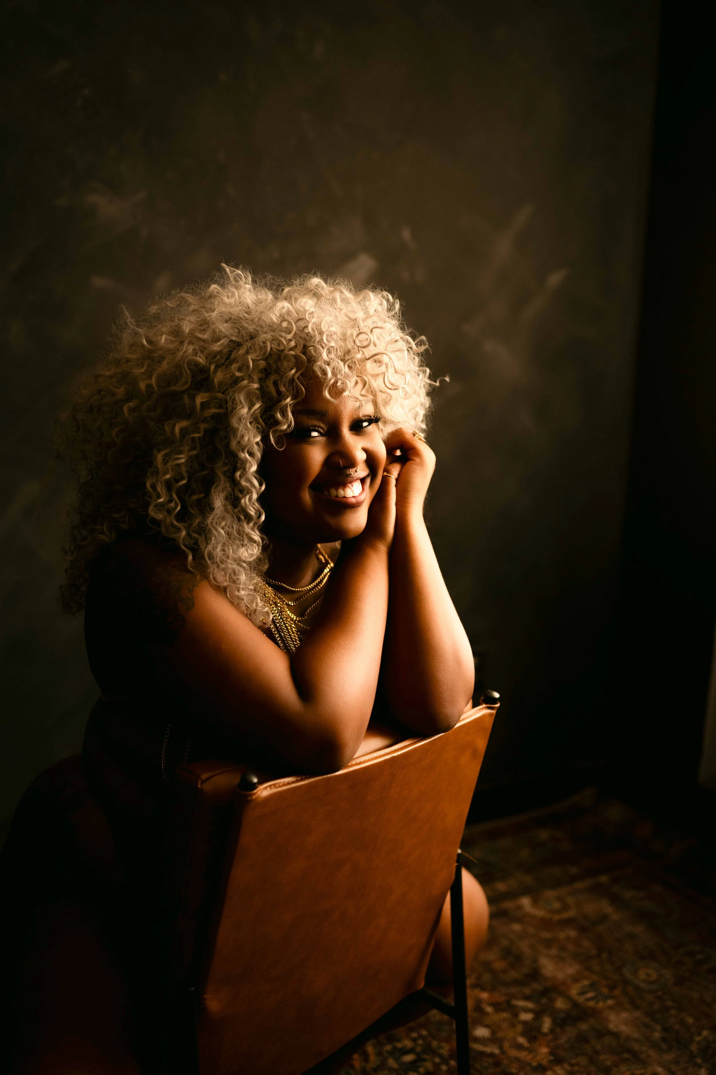 Smiling woman with curly blonde hair leaning over a chair during a warm, moody Houston boudoir session