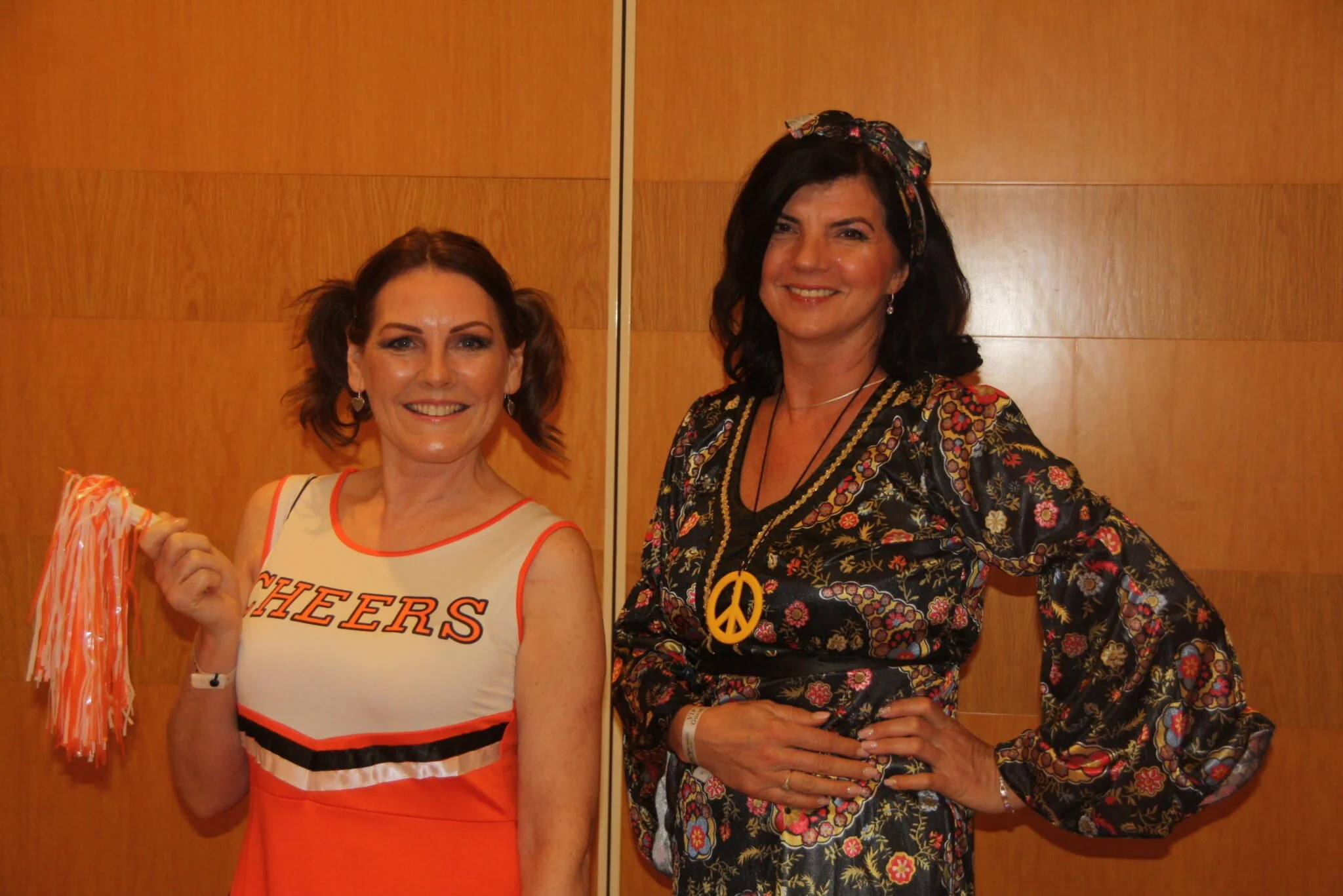 Two women standing indoors in front of a wooden wall, smiling. The woman on the left is dressed as a cheerleader holding pom-poms, while the woman on the right is wearing a floral dress with peace sign necklace.