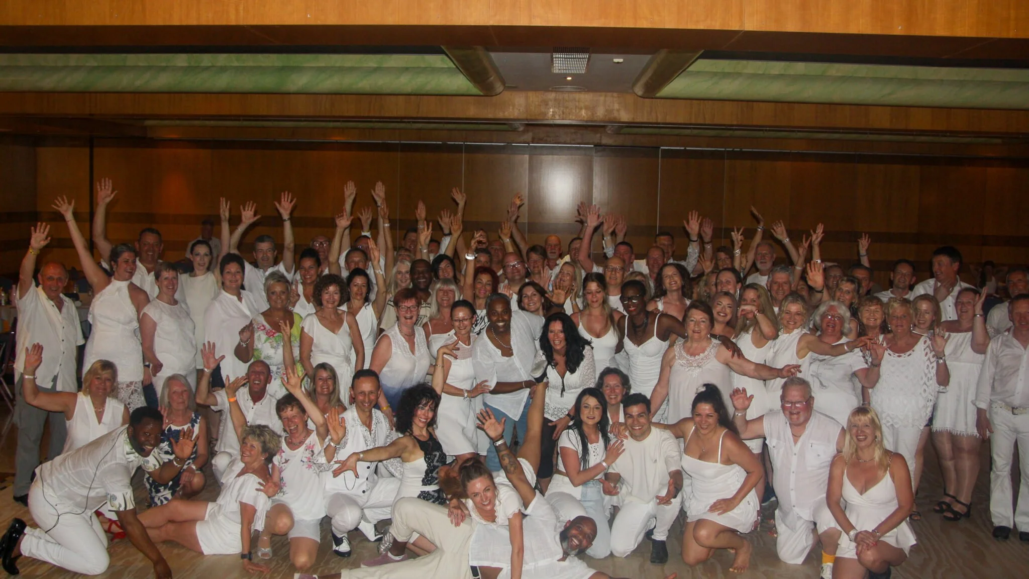 Large group of smiling people in white clothing gathered indoors, with many raising their hands.