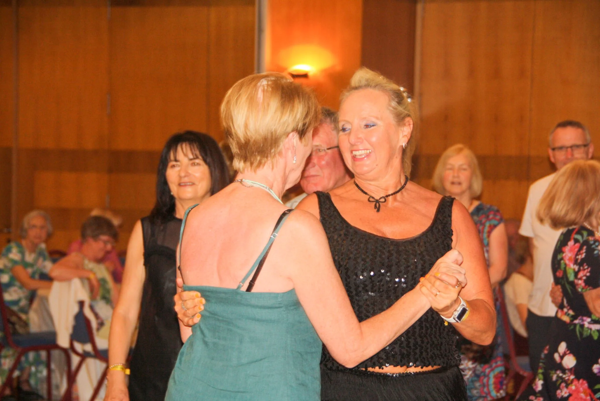 Two women dancing and smiling at a social gathering or party, with other people dancing and socializing in the background.