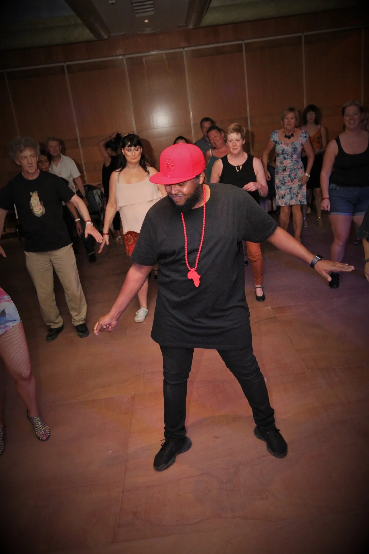 A man wearing a red cap, black t-shirt, and black pants is dancing with enthusiasm among a group of people in a room with wooden-paneled walls. The audience is watching, some smiling and others focused on dancing.