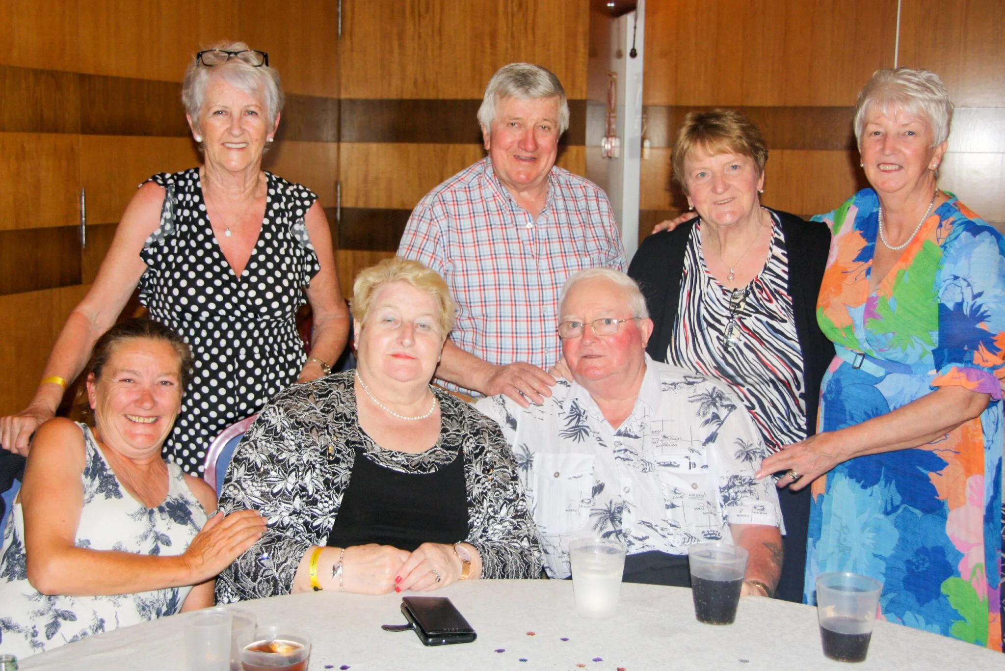 Group of nine elderly people, six women and three men, gathered at a social event indoors, smiling and posing for the photo, with drinks on a table in front of them.
