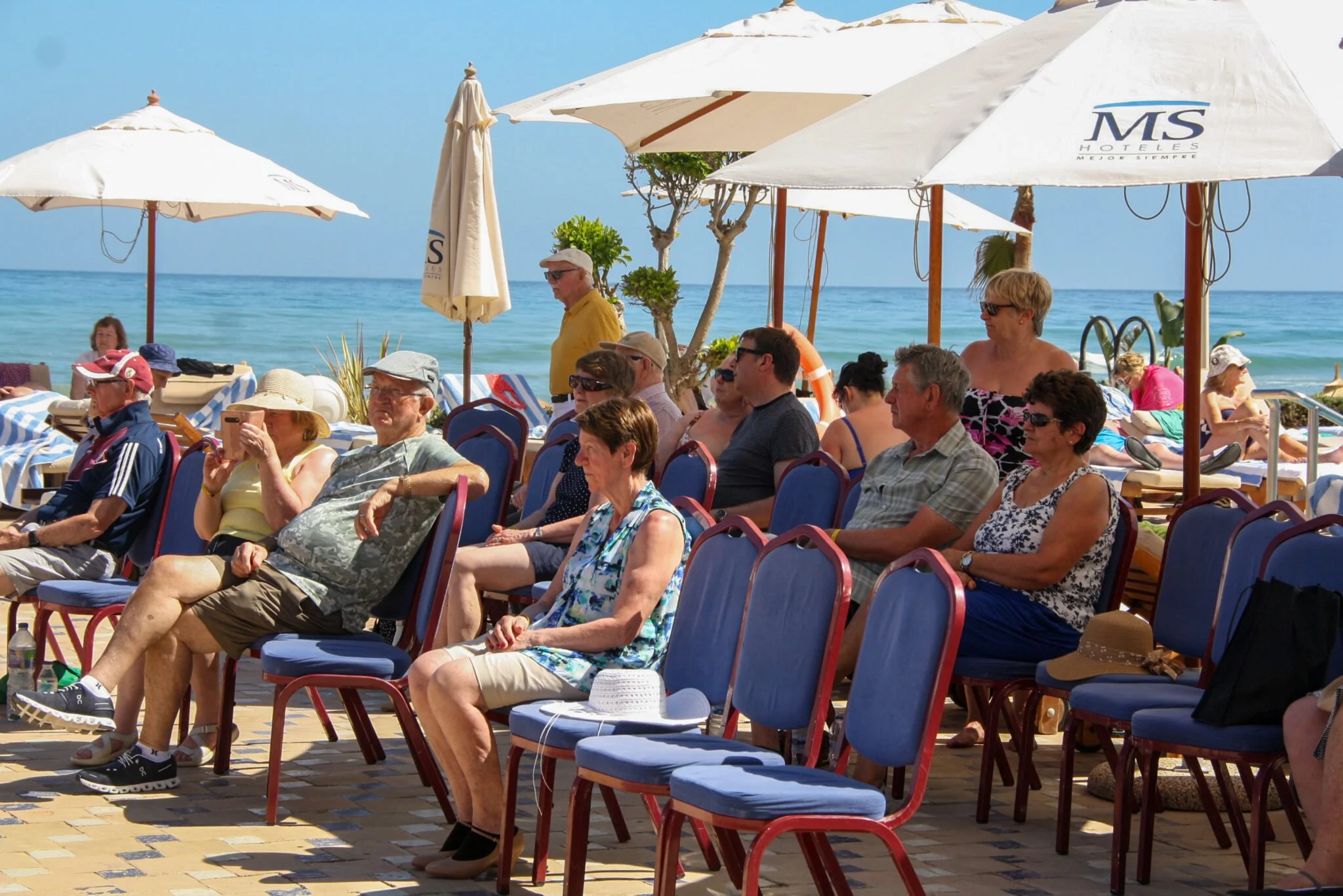 People sitting on chairs at an outdoor beachside event with umbrellas over looking the ocean.