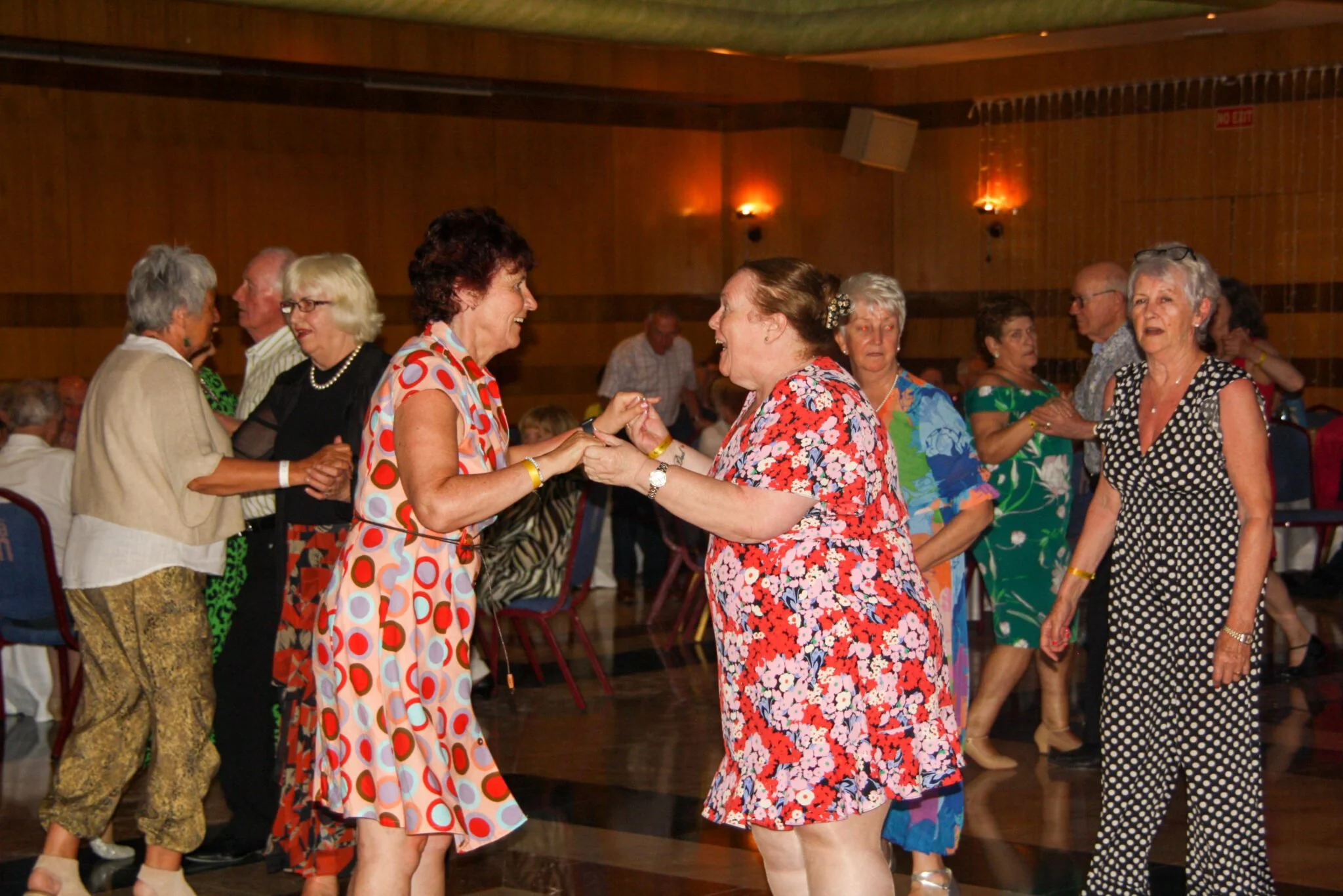 Group of elderly women dancing and socializing in a decorated wooden hall, dressed in colorful retro-style dresses.