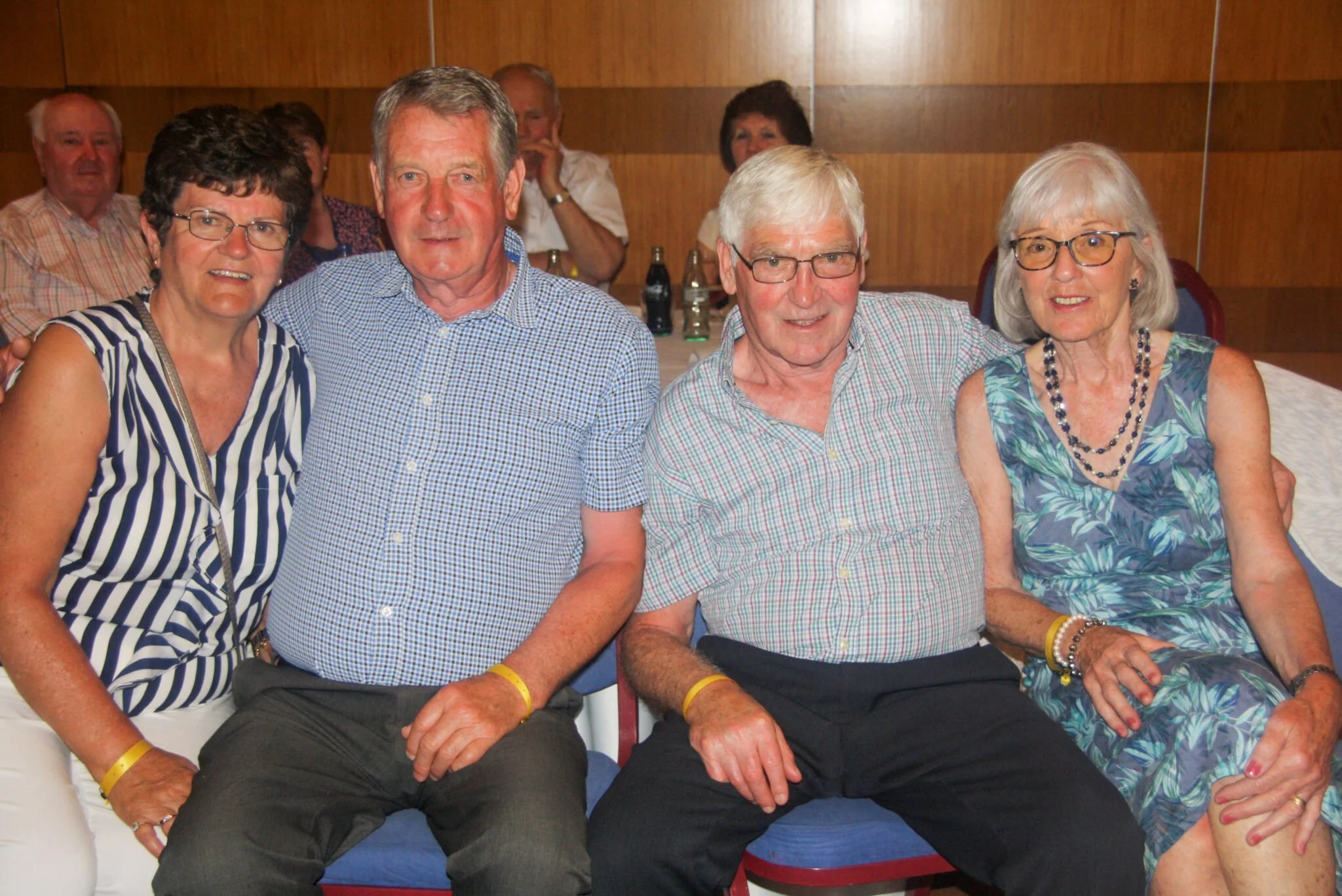 Four older adults sitting together, smiling, in a social gathering indoors. Two men are in the middle and two women are on the sides. They are wearing casual summer clothes.
