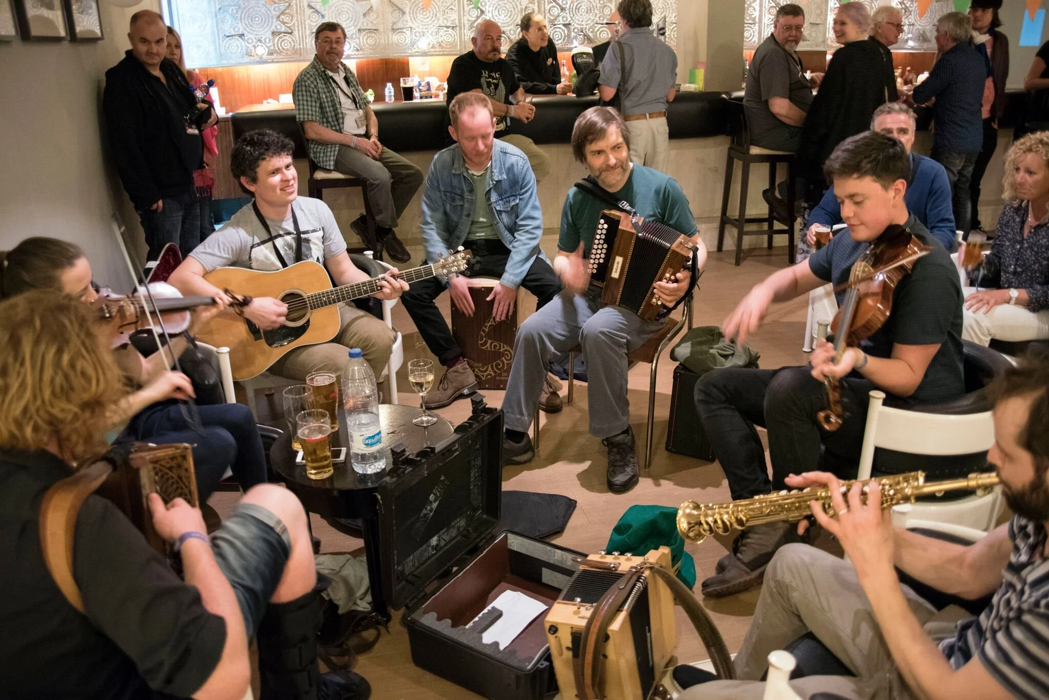 Group of musicians playing instruments in a social gathering, with people sitting and standing around, some holding drinks, in an indoor setting.