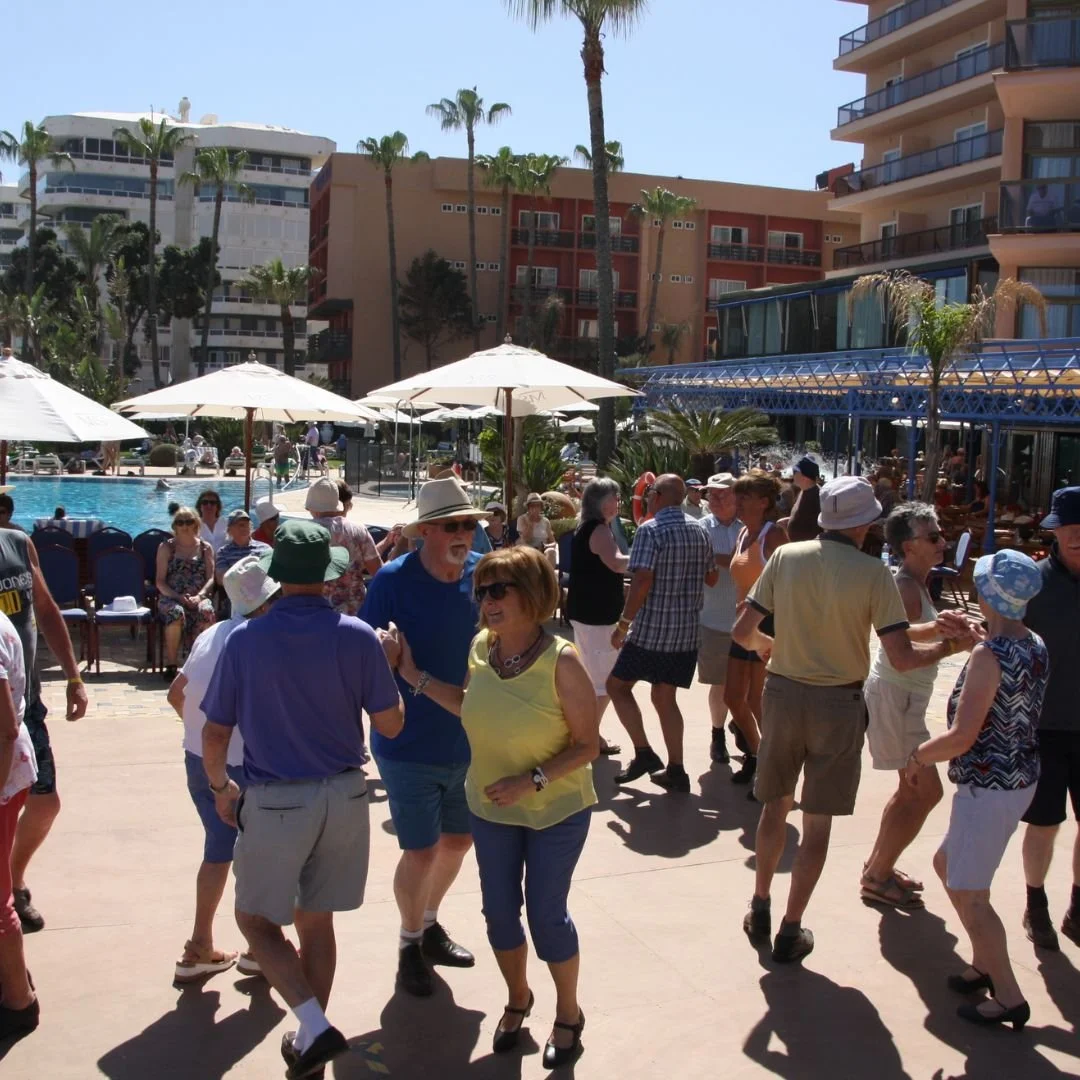 People dancing and socializing by a hotel poolside on a sunny day with palm trees and modern buildings in the background.