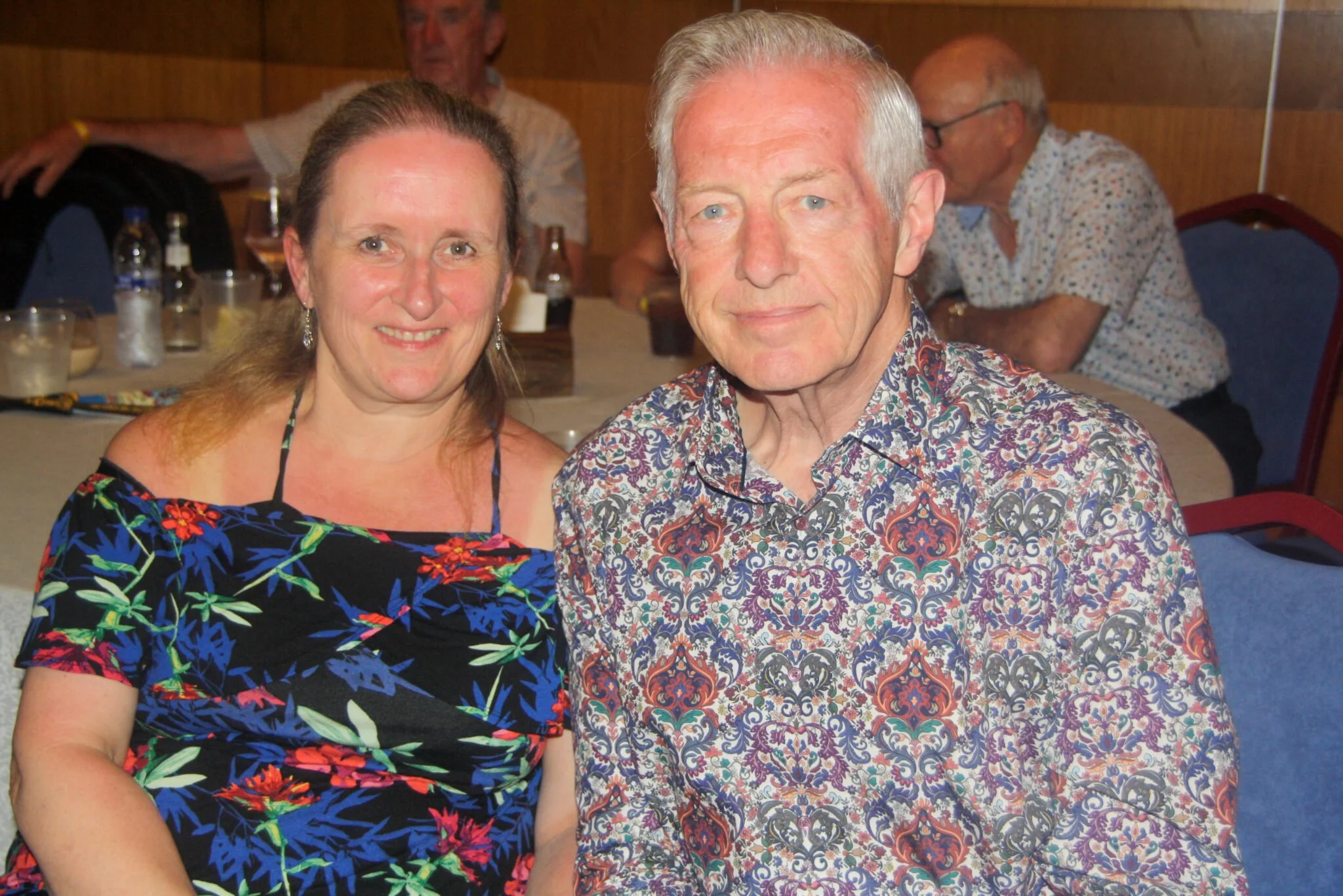 A smiling woman with long brown hair and earrings sitting next to an older man with gray hair wearing a patterned shirt, at a social gathering with other people in the background.