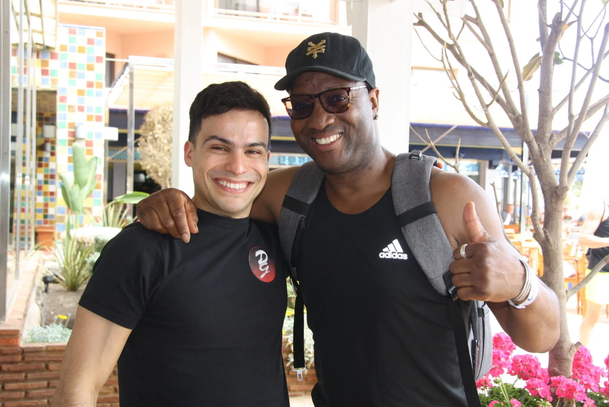 Two smiling men standing close together outdoors, one with his arm around the other's shoulder, giving a thumbs up with his hand. They are dressed casually, with backpacks, and seem happy. Background shows a tree, pink flowers, and a colorful mosaic