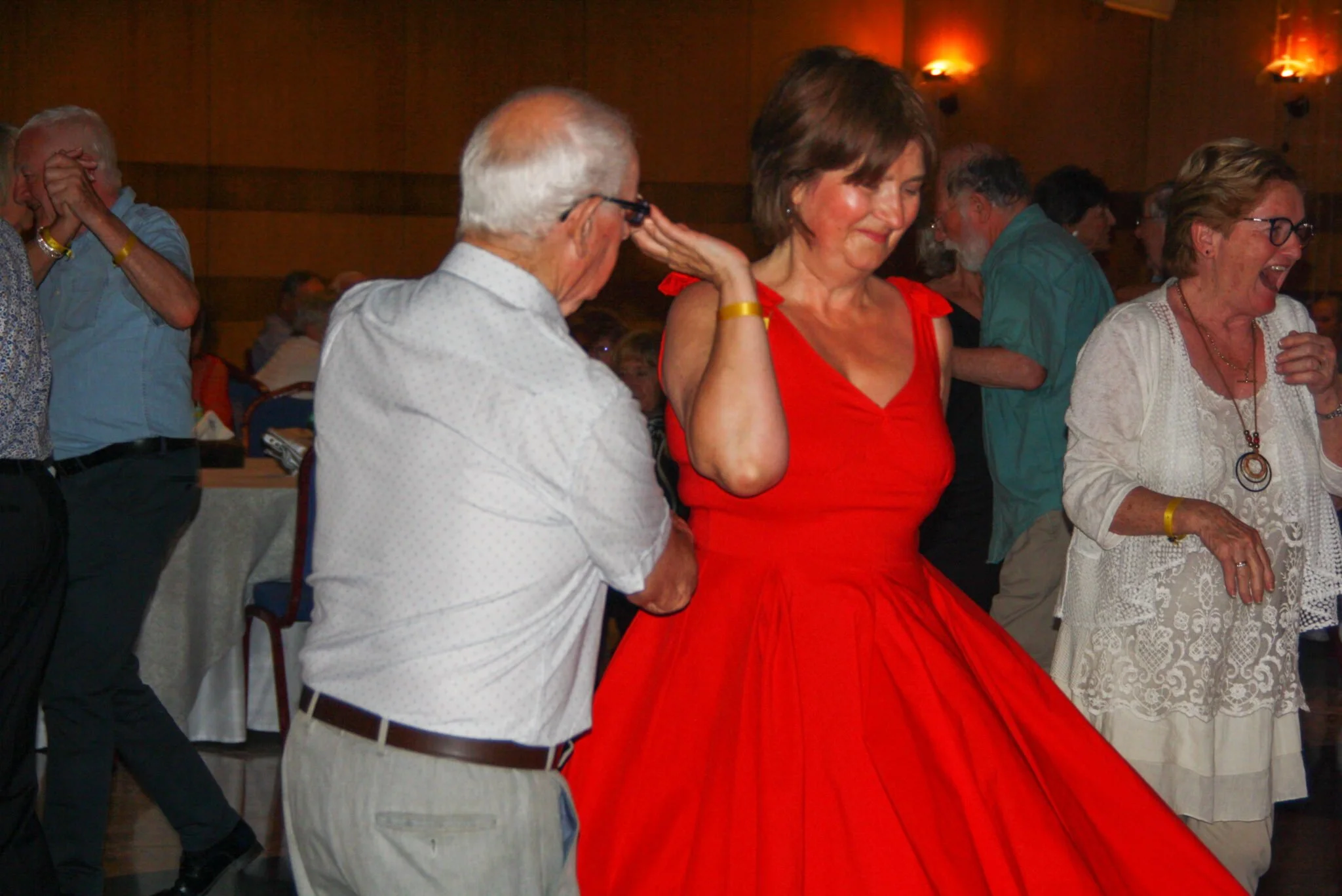Older adults dancing at a social event, with a woman in a red dress in the center and others around her, in a warmly lit room.