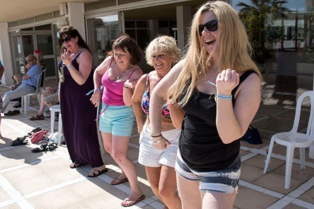Group of five women standing on a sidewalk near a pool, smiling and holding each other's hips in a line, with chairs and other people in the background.