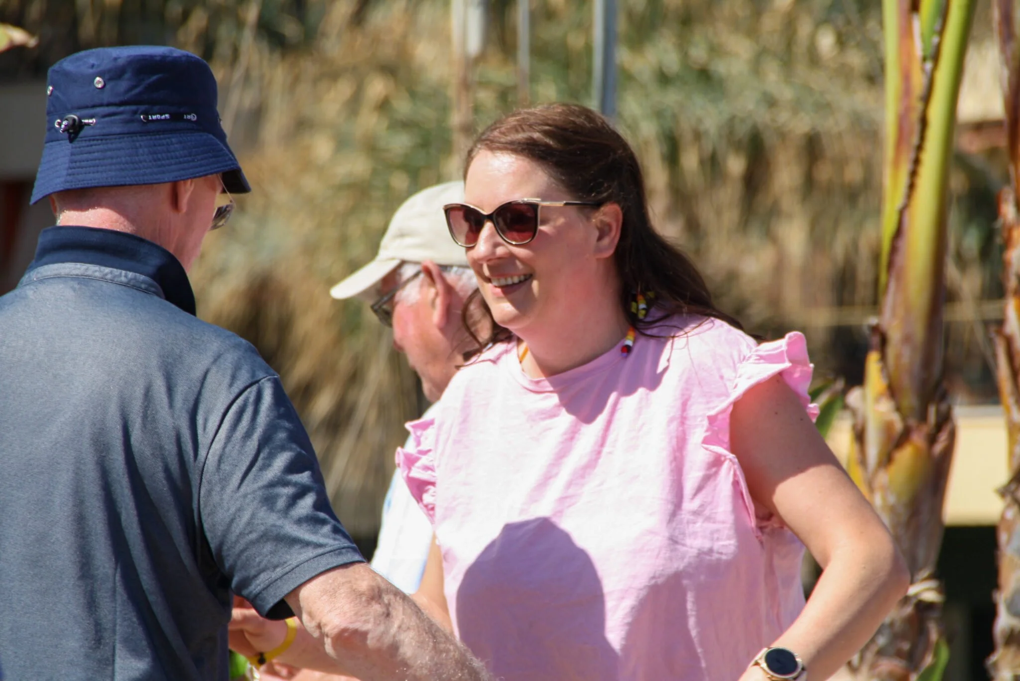 Group of people outdoors, woman wearing sunglasses and pink ruffled top smiling and talking to a man in a blue hat and shirt.