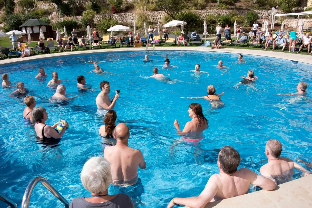 People swimming and relaxing in a large outdoor swimming pool, with others sitting on lounge chairs and umbrellas around the poolside.