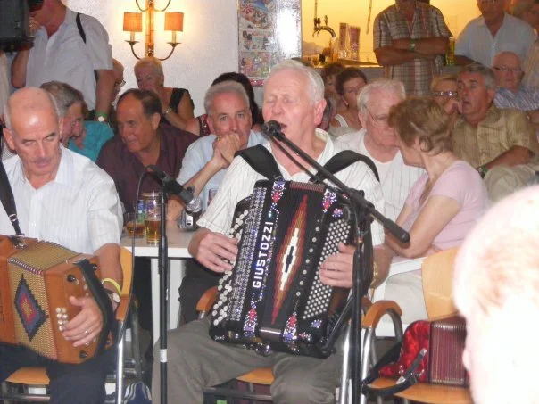 A man playing an accordion at a well-attended Gerry Flynn event, surrounded by seated attendees.