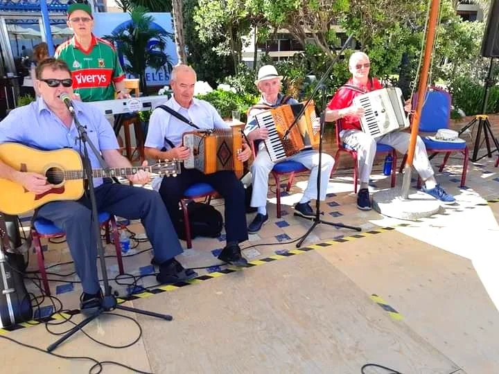 Group of five men performing music outdoors with guitars and accordions, one man standing behind them playing keyboard in the background.