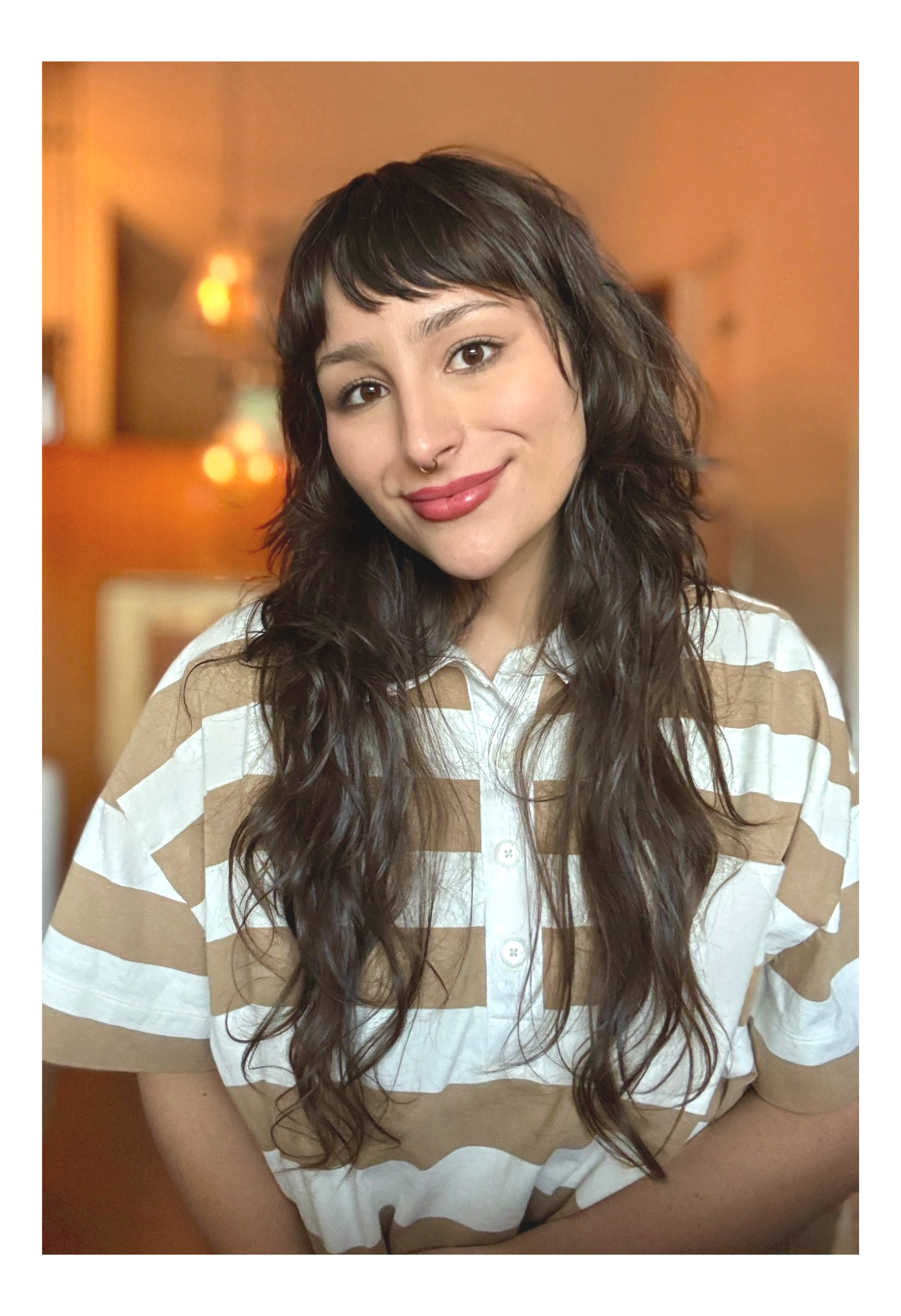 A young woman therapist with dark wavy hair and a septum piercing smiling at camera, wearing a striped beige and white shirt, in a warmly lit indoor setting.