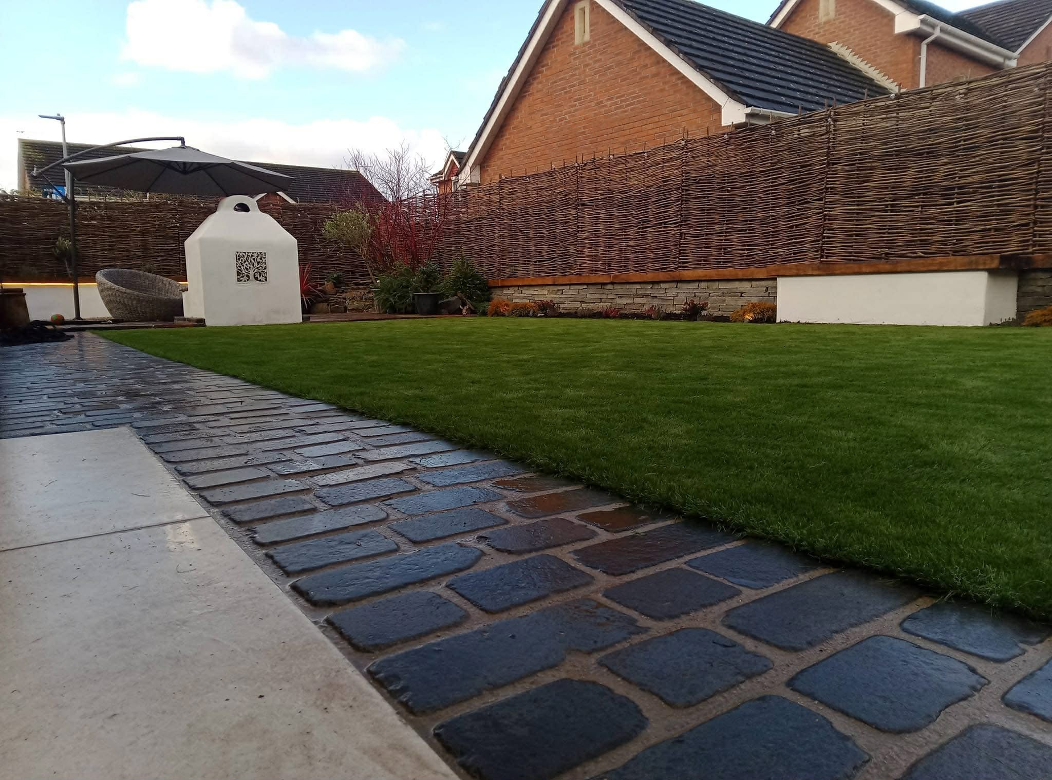 Low maintenance garden design and installation near Crewe with porcelain paving, artificial grass installed plus hazel hurdle fencing, granite cobble pathway, outdoor fireplace and much more!
