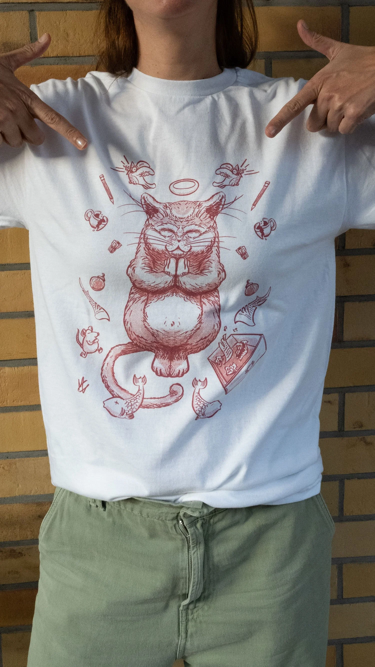 Person wearing a white t-shirt with a drawing of a cat praying and surrounded by fish, mice, and game pieces, pointing to the design with both hands, standing against a brick wall.