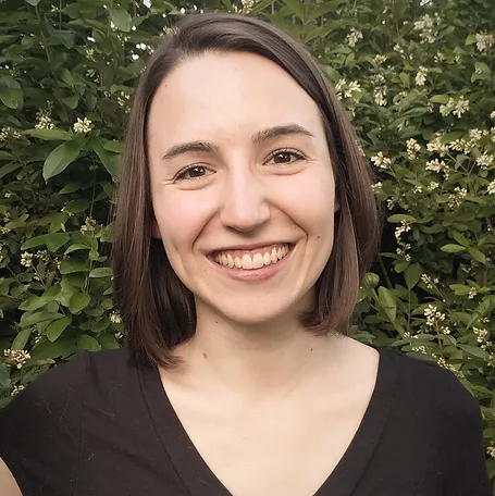 A woman with shoulder-length dark hair smiling outdoors in front of green leafy bushes.