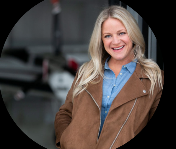 A woman with blonde hair, smiling, wearing a blue shirt and brown jacket, standing in front of a blurred background with cars.