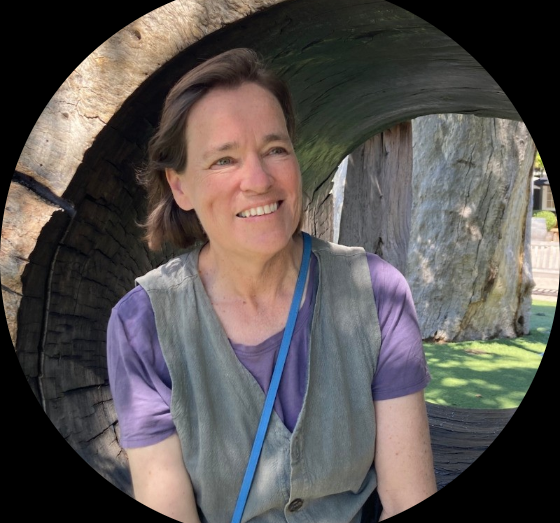A woman with short brown hair and a purple shirt smiling outdoors, sitting inside a large hollowed-out tree trunk.