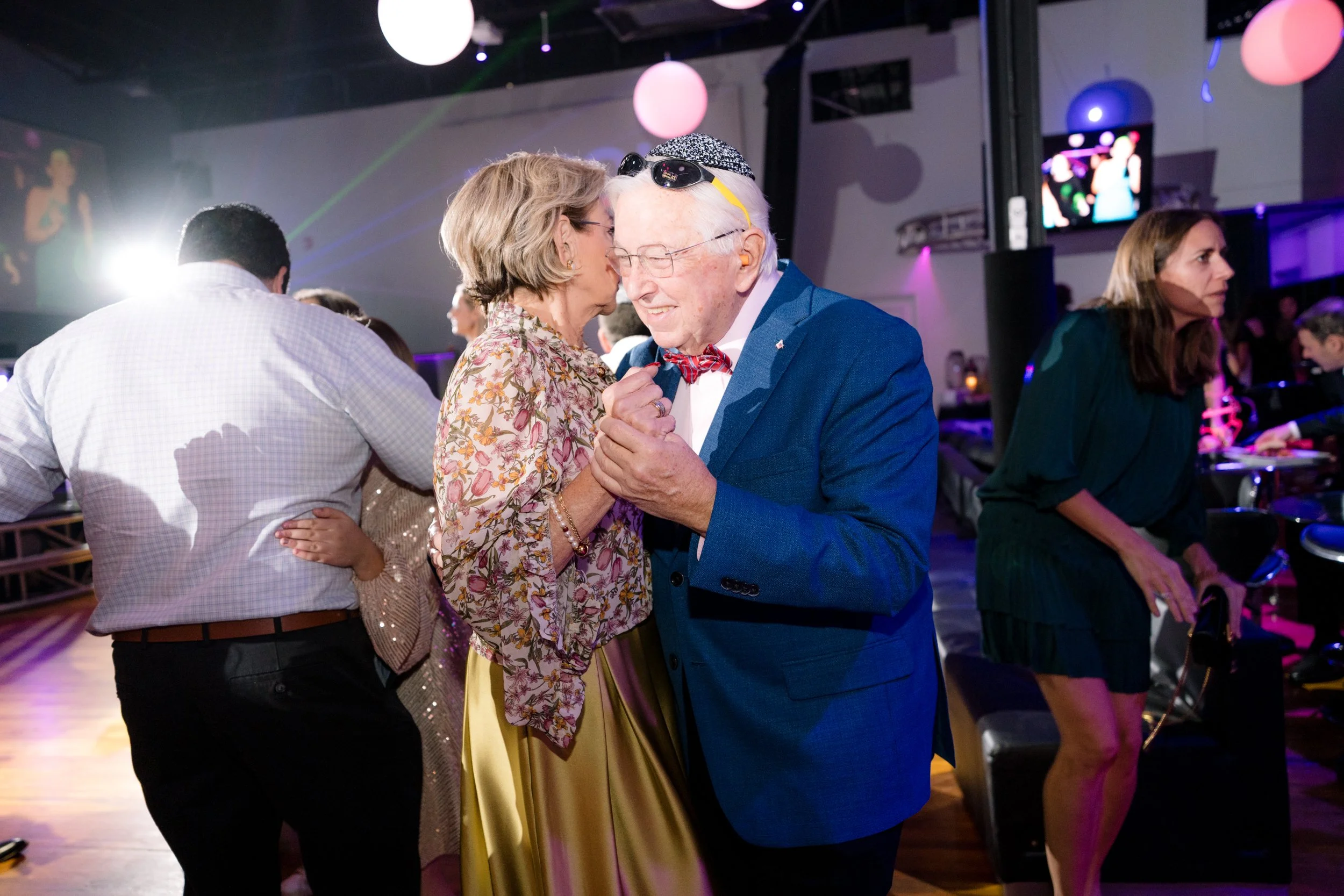 Elderly couple dancing at a party with ambient lighting and people socializing in the background.