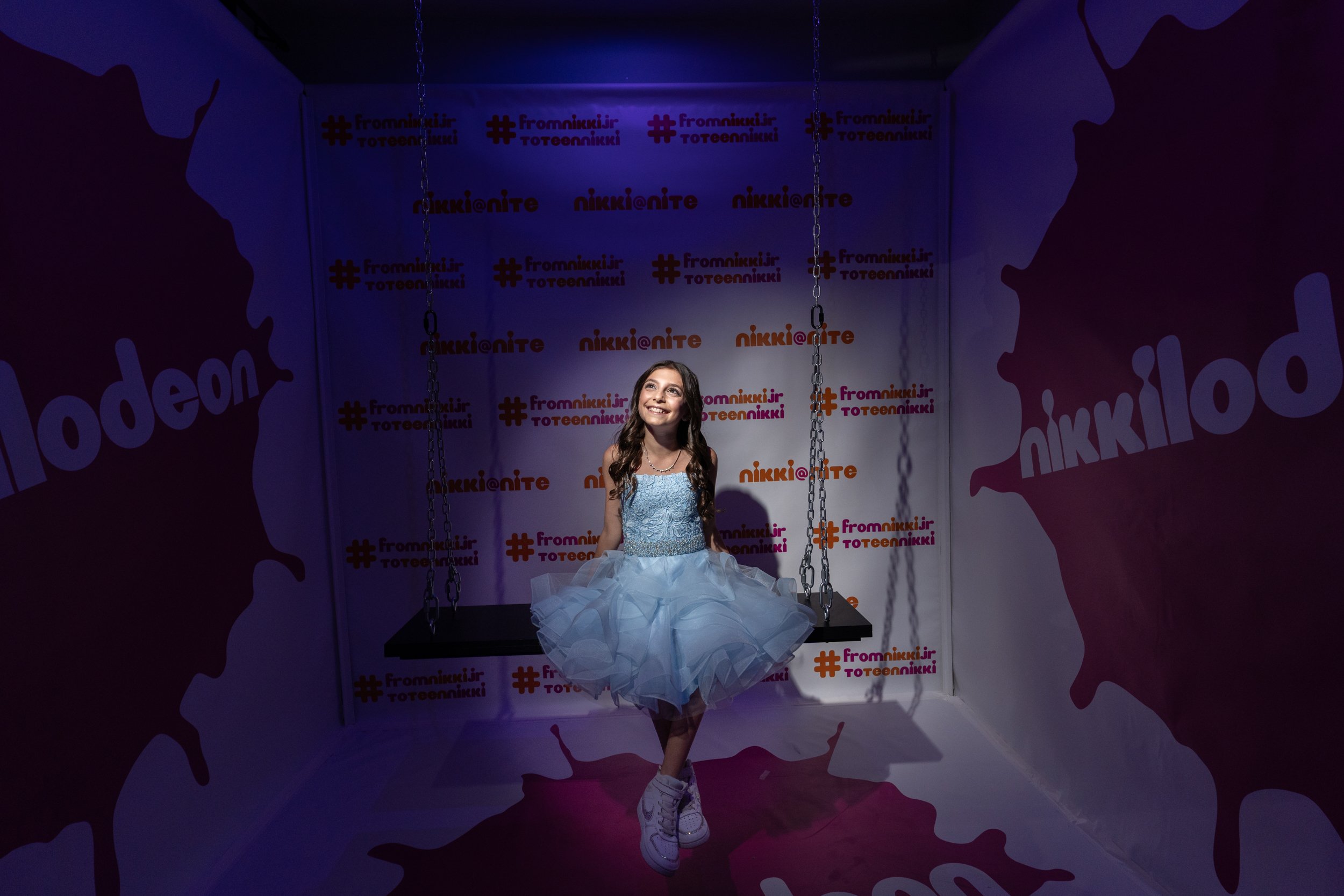 Girl in blue dress sitting on a swing in a colorful Nickelodeon-themed photo booth with hashtags and logos on the walls.