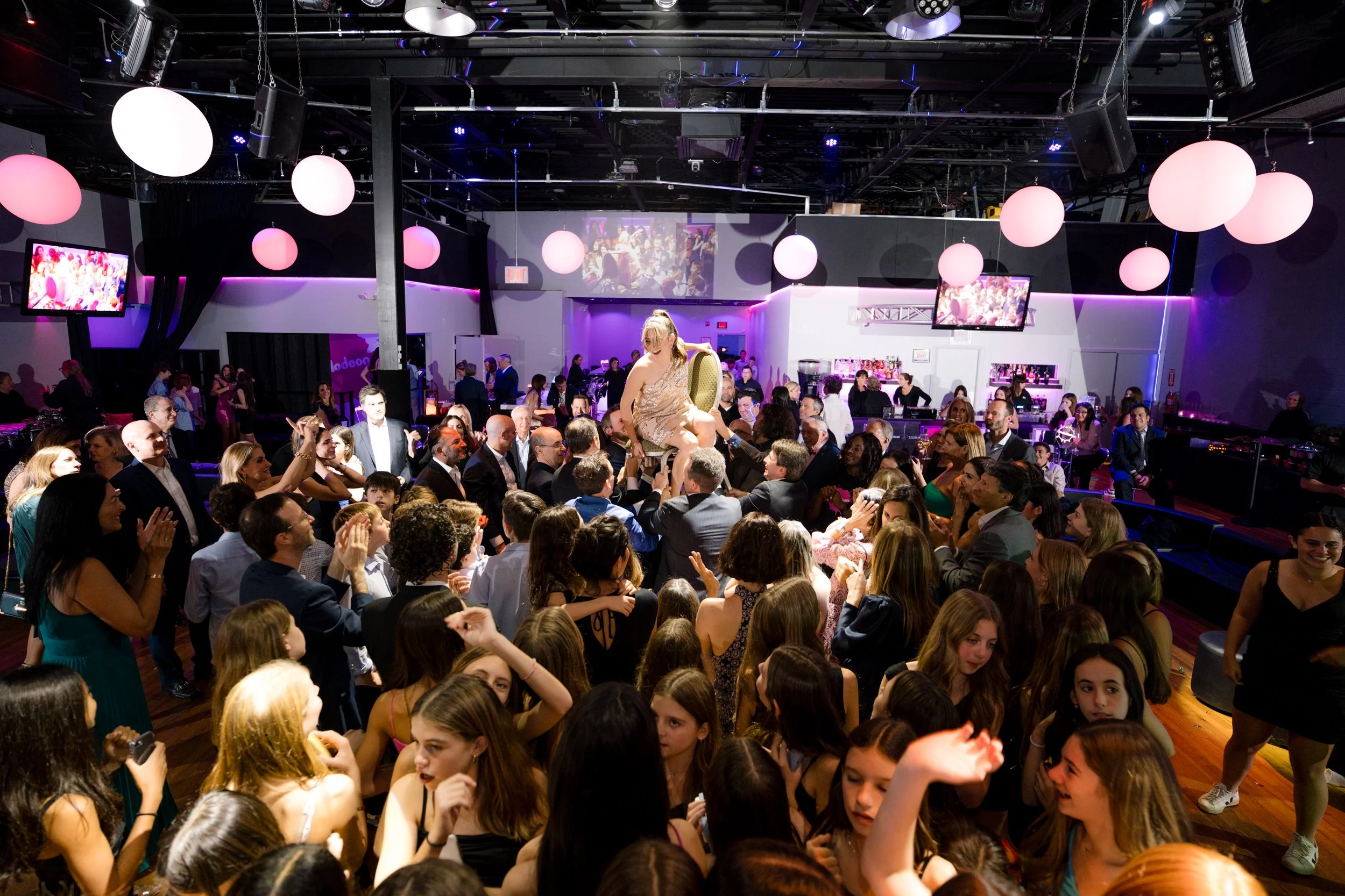 A group of people at a lively indoor event, featuring a person being lifted on a chair in the center of the room, surrounded by a crowd. The venue is decorated with hanging spherical lights and has a bar in the background. The atmosphere is festive.