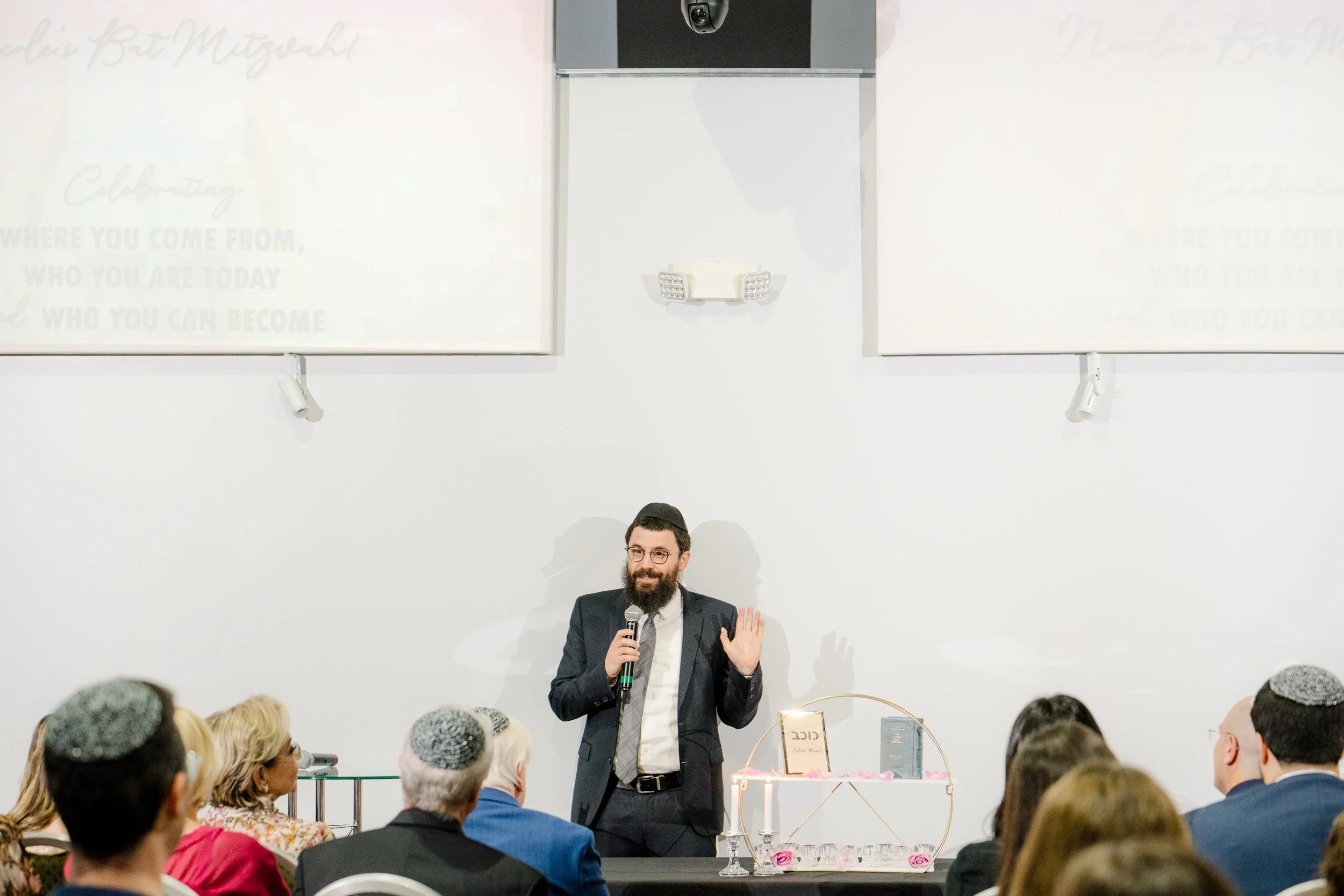 A man in a suit speaking into a microphone at an event, standing in front of a table with books and candles. Attendees are seated, wearing kippahs.