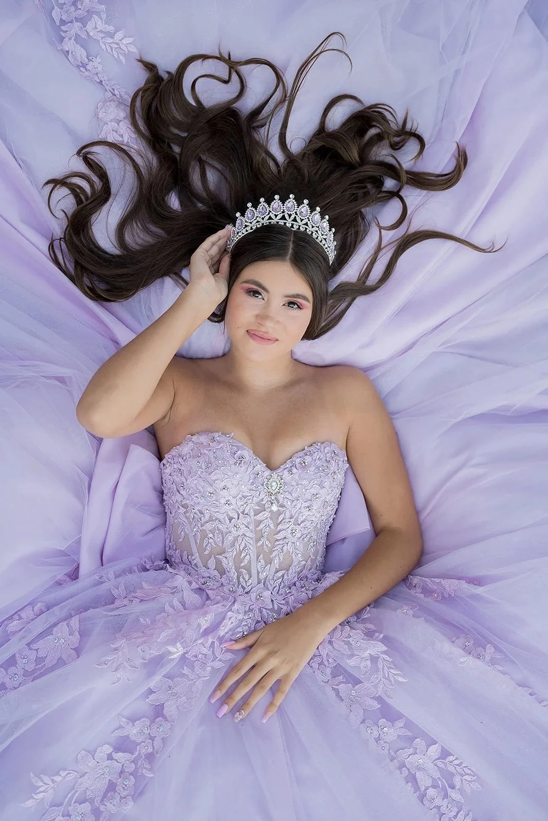 A quinceañera girl with long, wavy hair lying on a lavender fabric, wearing a lavender strapless dress with lace and floral embroidery, and a silver tiara on her head.