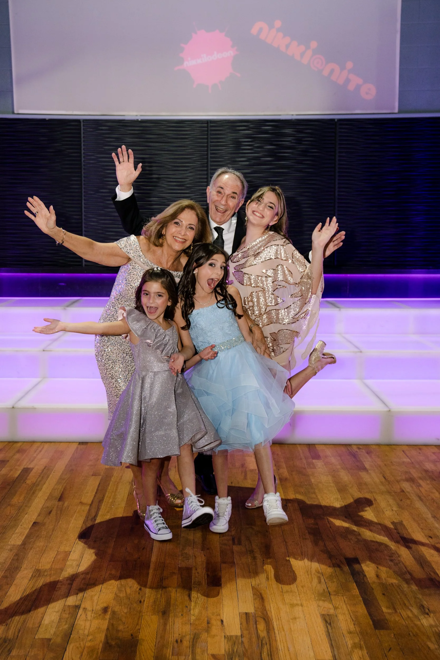 A group of people posing happily, including young girls in party dresses and sneakers, alongside adults in formal attire, against a festive backdrop with colorful lights.