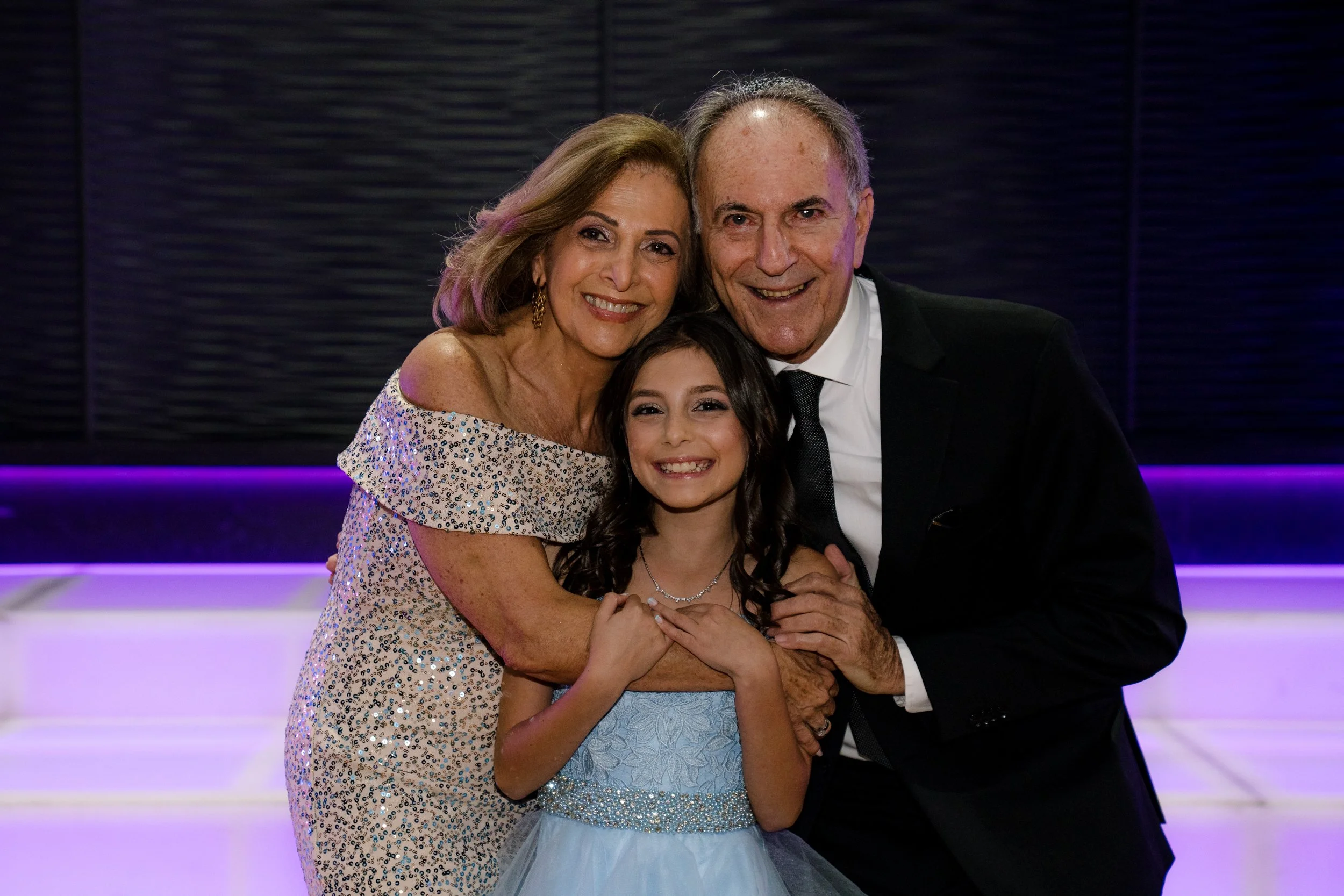 A joyful family portrait featuring an elderly couple and a young girl, smiling together in formal attire against a purple-lit background.