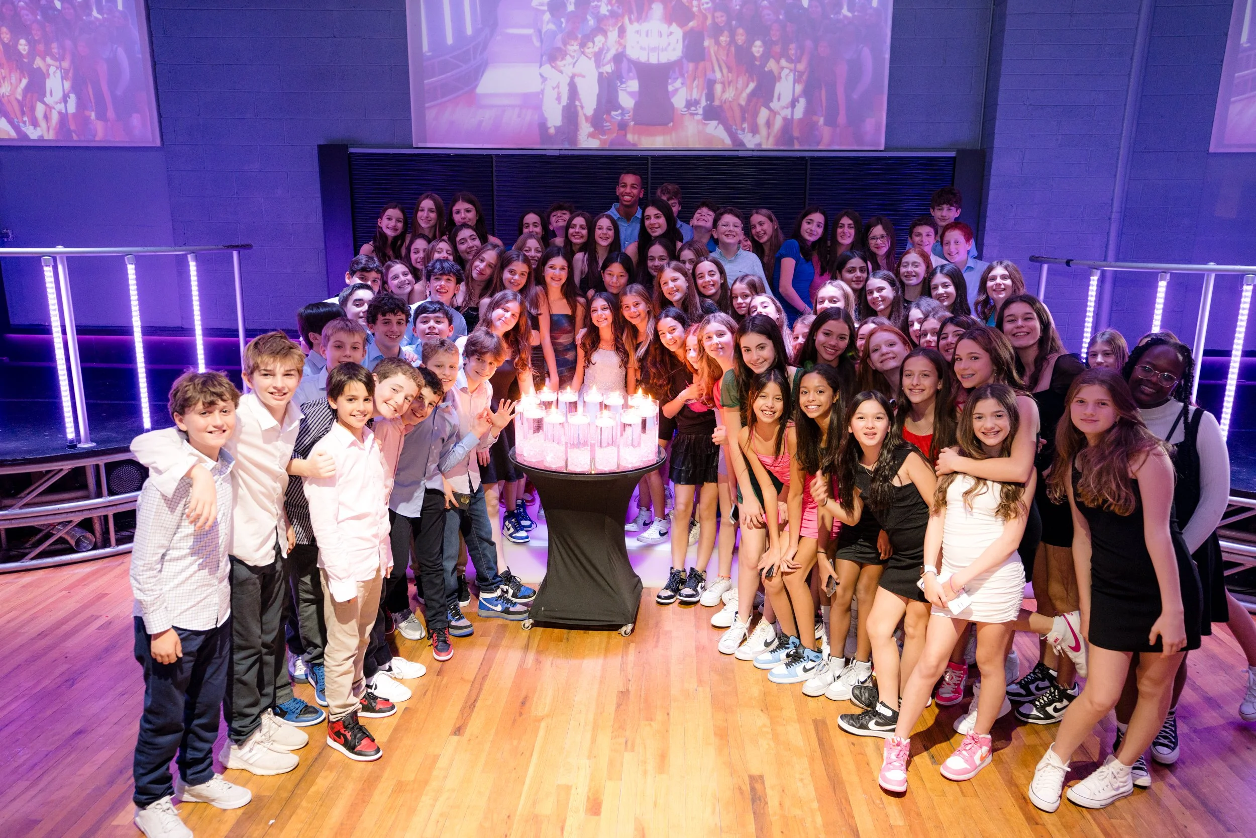 A large group of young people gathered around a table with lit candles, celebrating at an event in a decorated venue.
