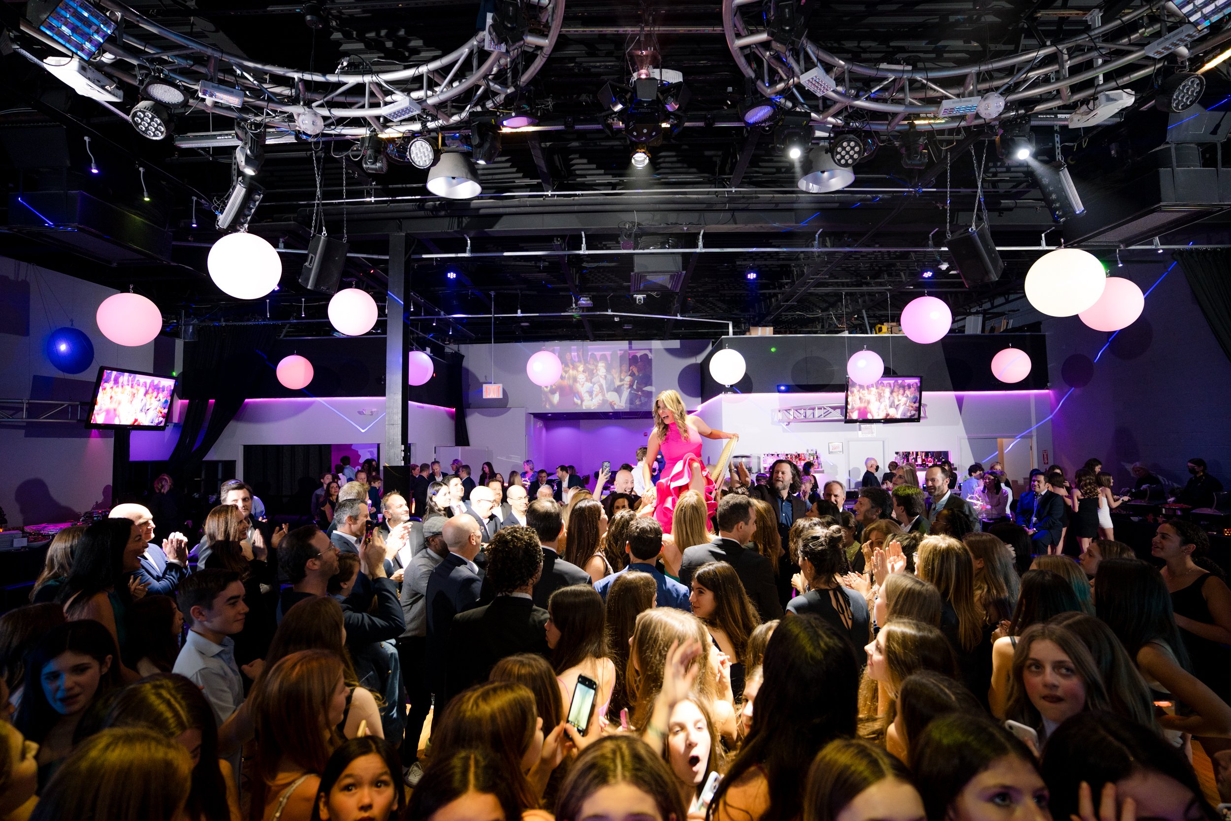 A crowded party with people dancing and celebrating under colorful lighting and round hanging decorations.