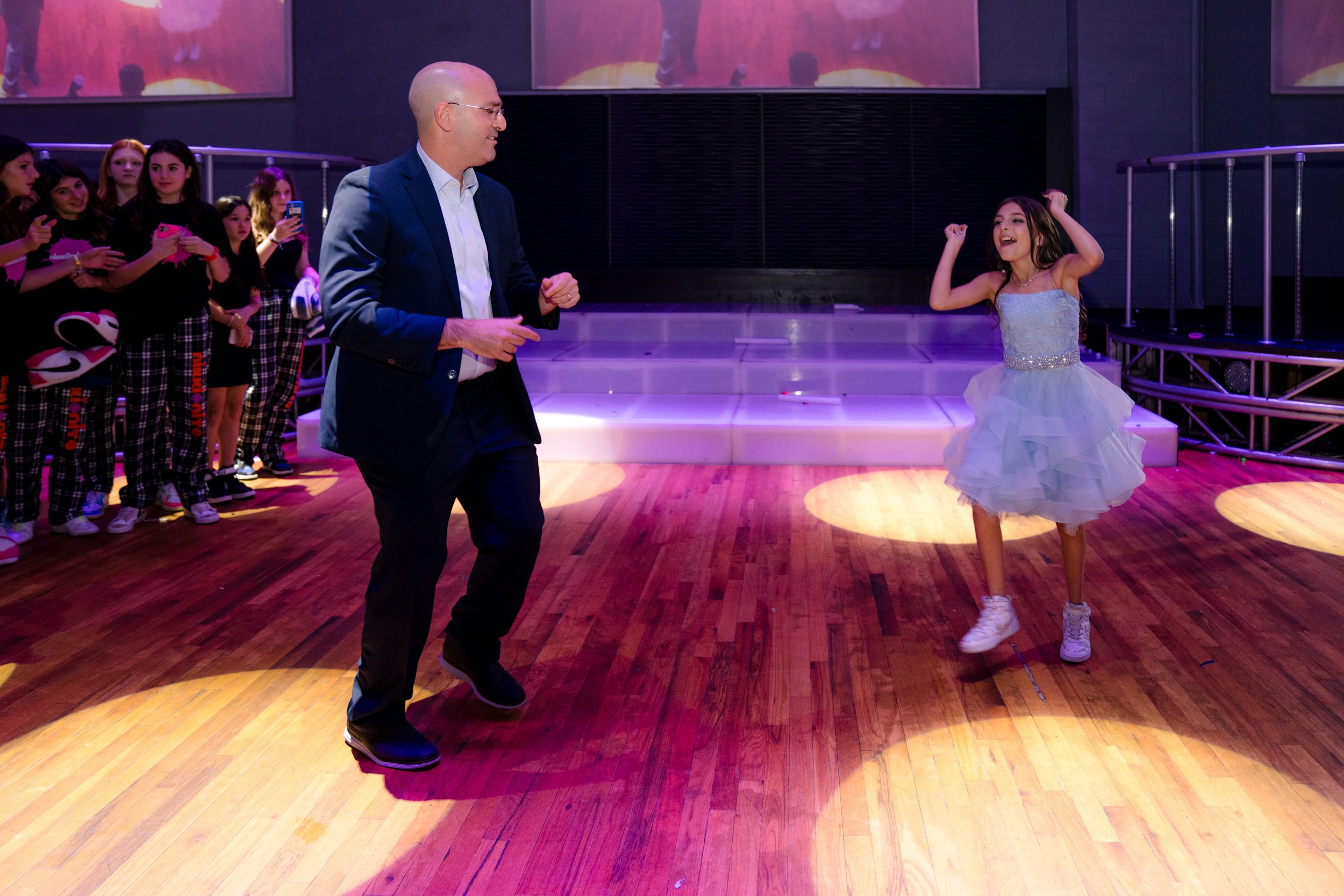 A man and a young girl dancing at an event, surrounded by people watching. The man is in a suit, and the girl is wearing a blue dress and sneakers. The venue has wooden floors and stage lighting.