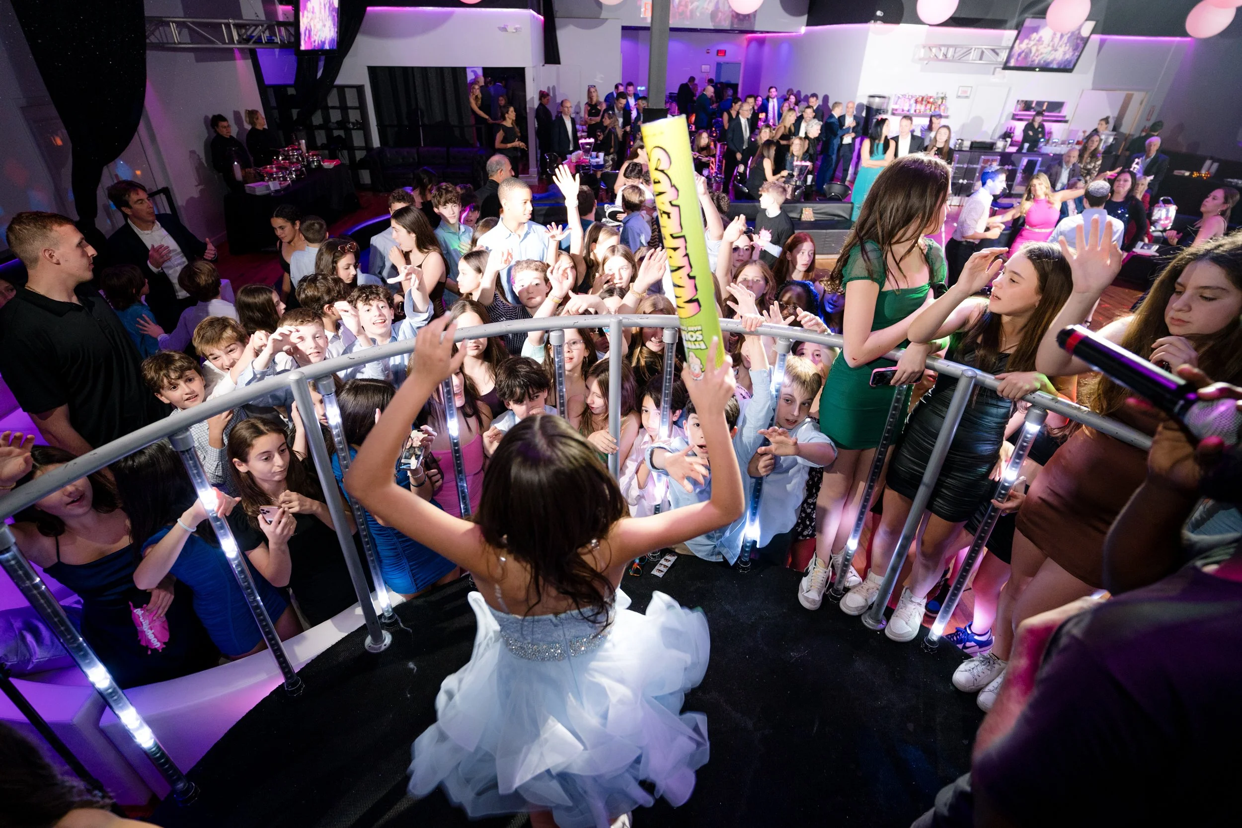 A lively party scene with a young girl wearing a blue dress, holding a large candy item, stands on a raised platform surrounded by a crowd of enthusiastic kids and teenagers. The atmosphere is festive with pink and purple lighting, and there are adul