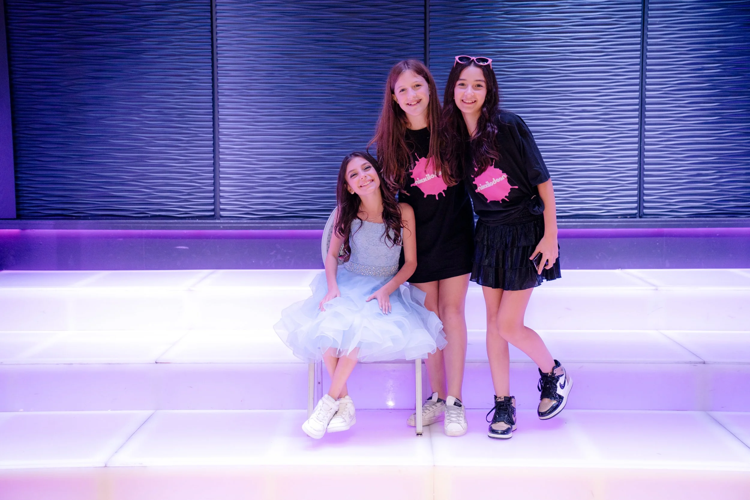 Three girls posing on an illuminated staircase, with one sitting in a blue dress and white sneakers, and two standing, wearing black t-shirts and skirts.