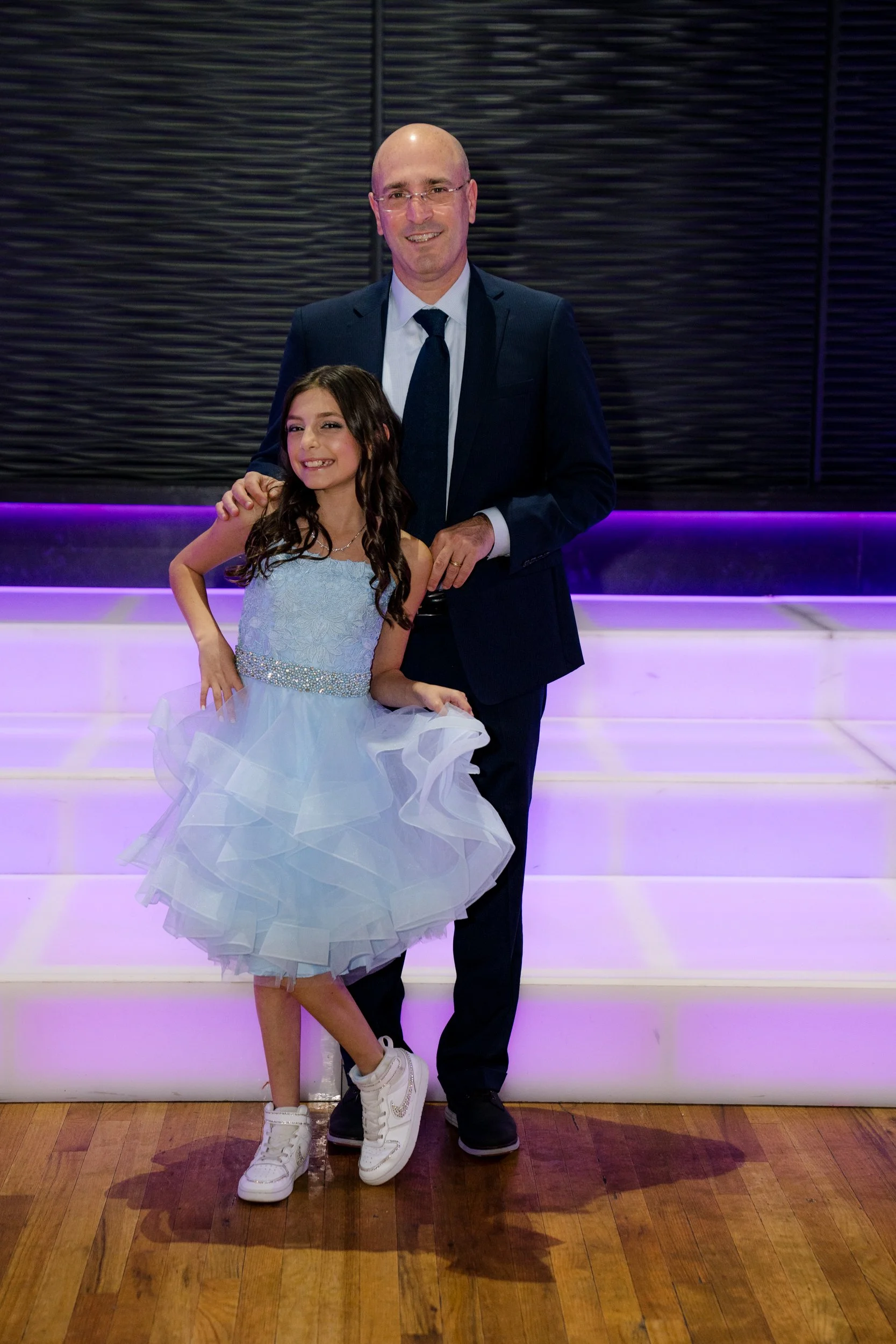 A young girl in a light blue dress stands beside a man in a suit, posing on a lit-up staircase with a dark background.