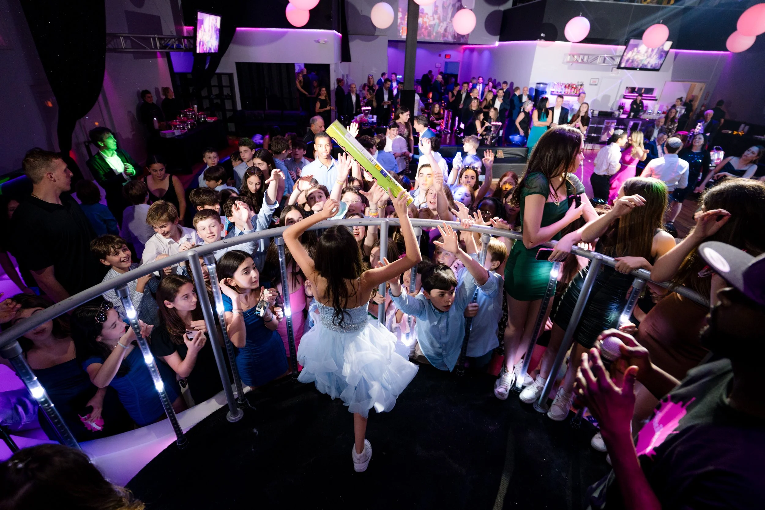 A lively party scene featuring a crowd of people, including many children, gathered around a girl in a blue dress holding a large inflatable stick. The venue is decorated with pink and purple lighting, with additional guests in the background near a 