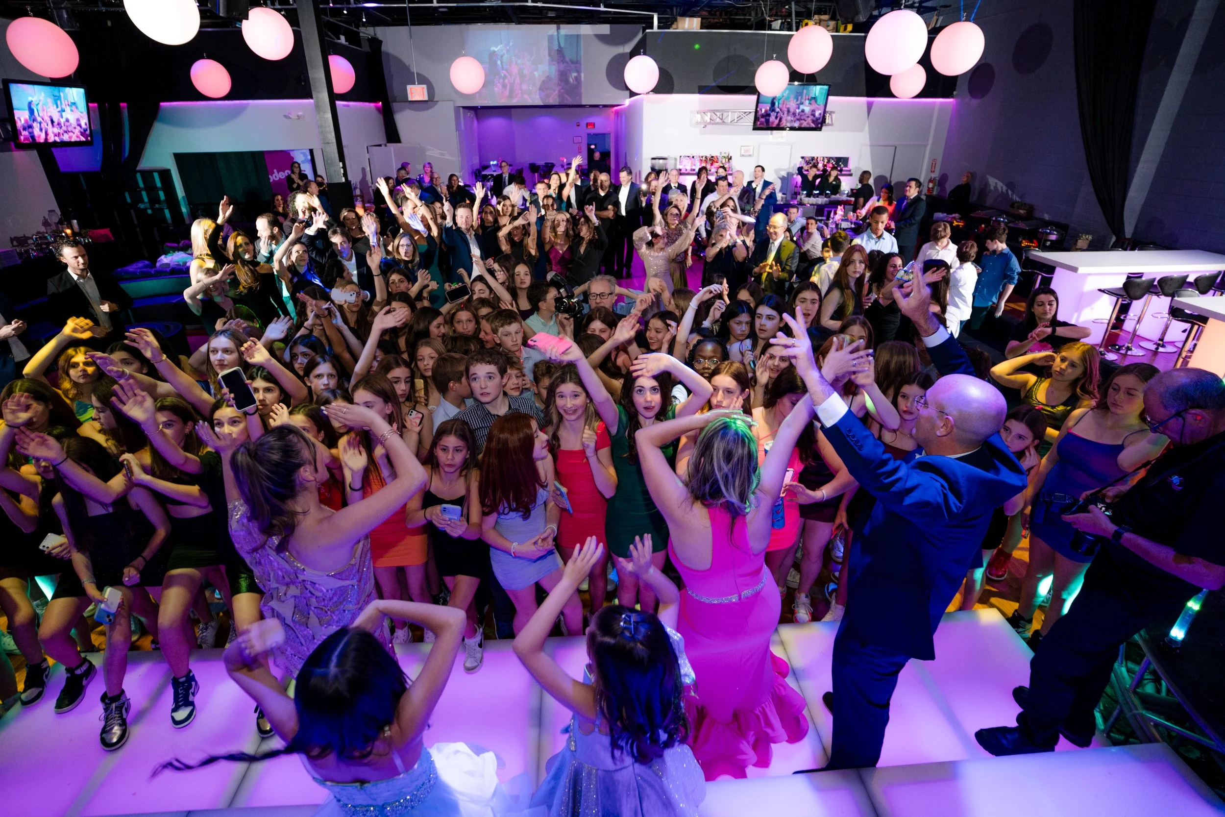 Crowd of people dancing at a party in a brightly lit venue, with colorful balloons and a stage area.