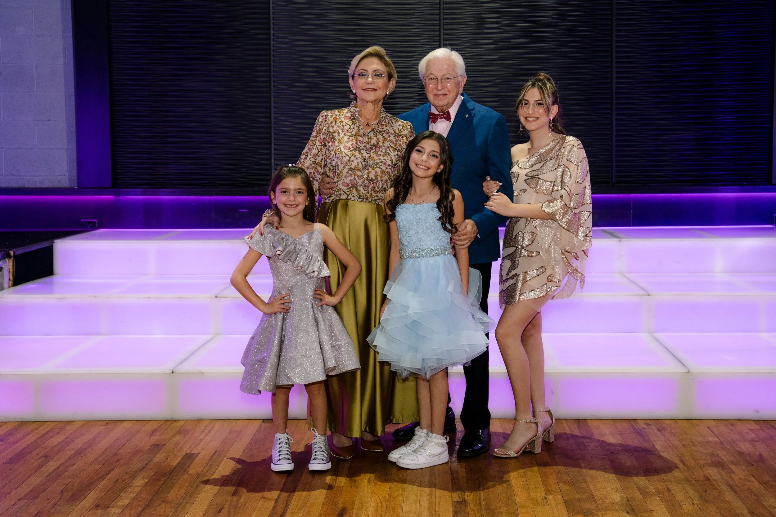 Group portrait of five people, including two adults and three children, standing on a lit stage with a wooden floor and purple lighting in the background.