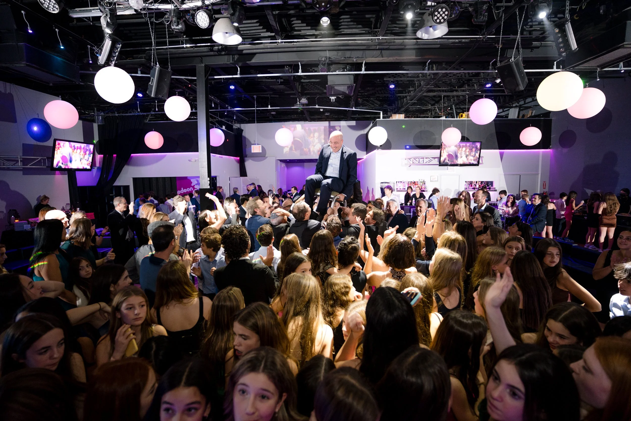 A lively indoor celebration with people dancing and a man being lifted on a chair. The room has colorful lighting and decorative hanging globe lights.